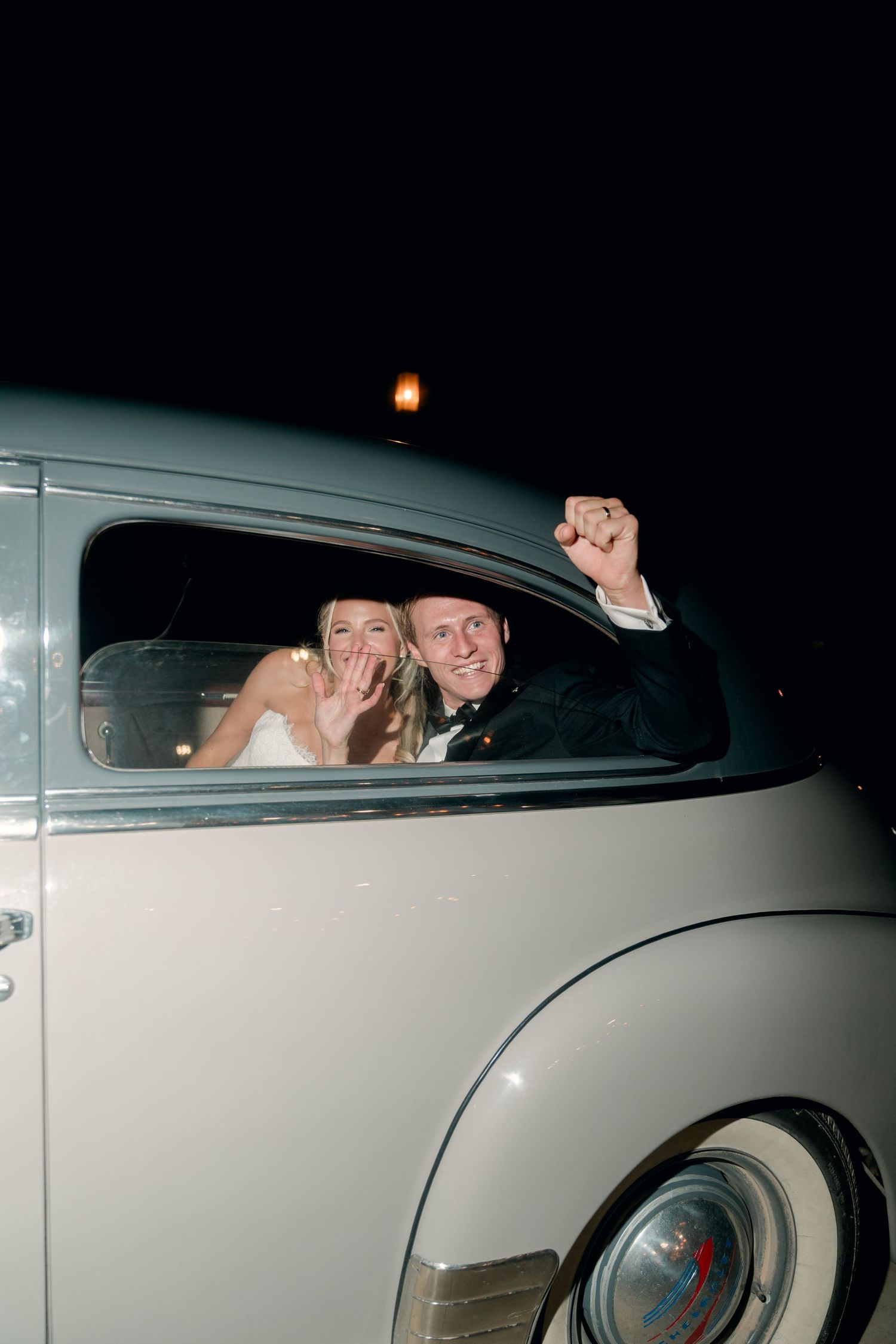 Newlywed couple celebrating in a vintage white classic car at night, bride smiling and groom raising fist in joy.