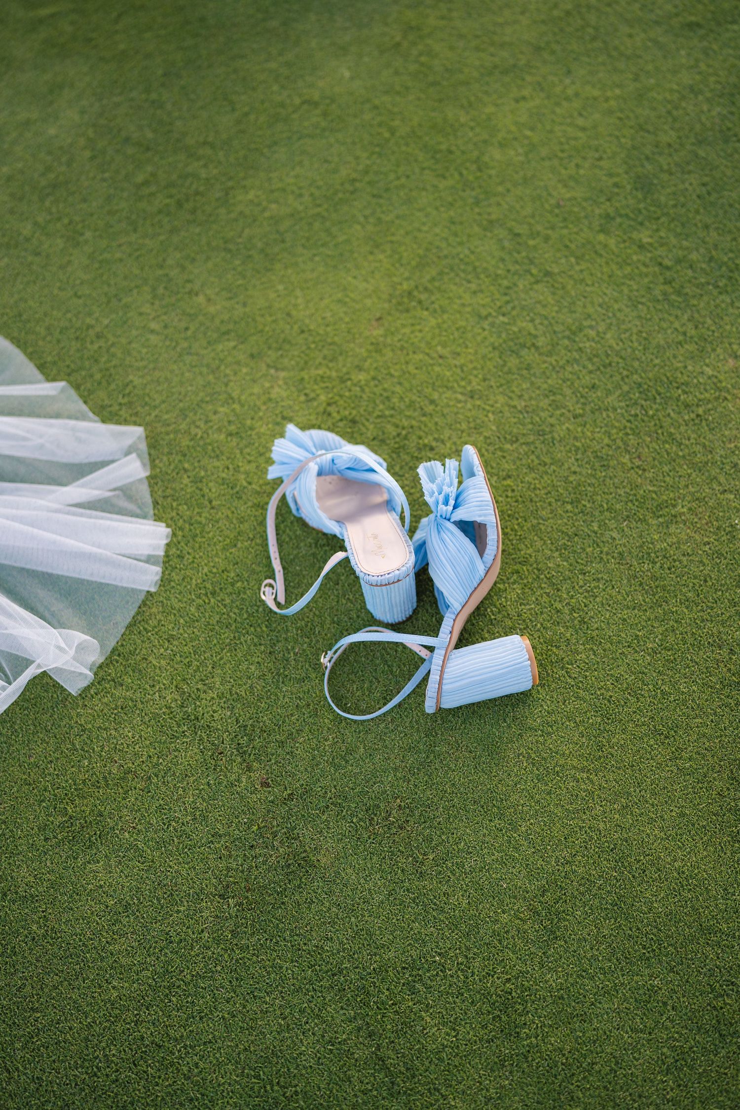 Light blue wedding shoes with delicate bows rest on green grass.