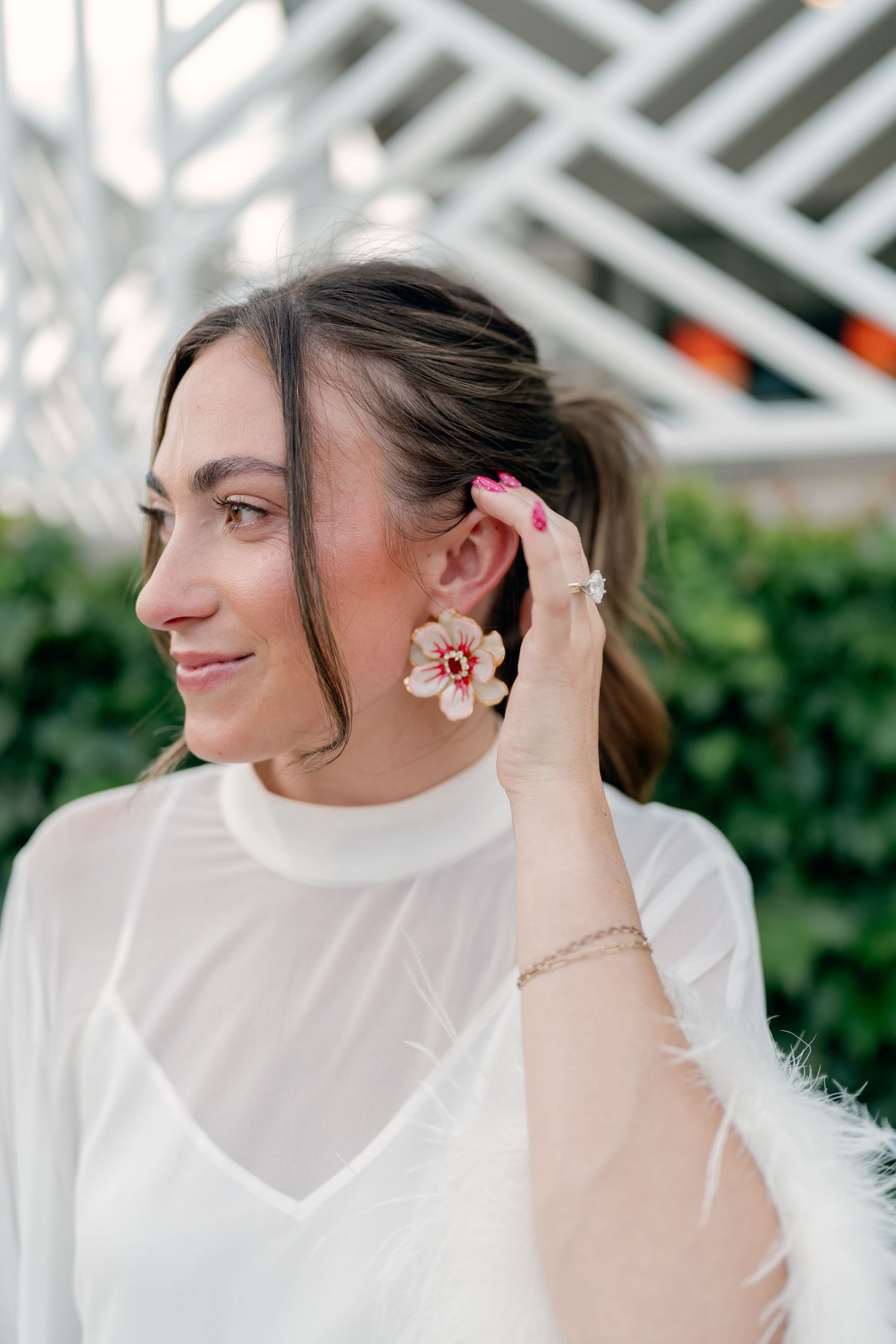 Bride in white dress showcases floral drop earrings while posing outdoors before wedding ceremony.