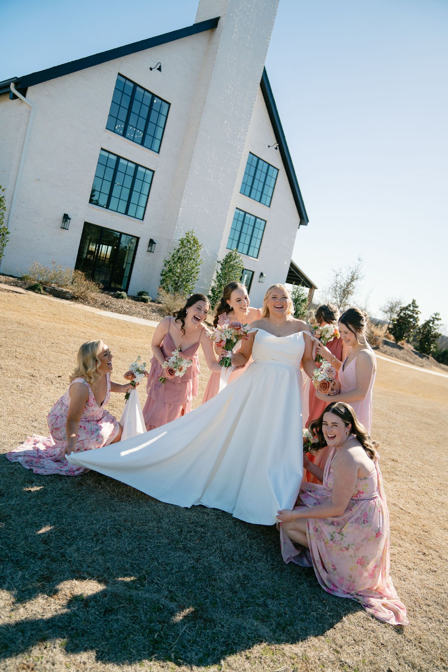 Bridesmaids in pink dresses sit together on the ground with the bride in a white gown during an outdoor photoshoot.