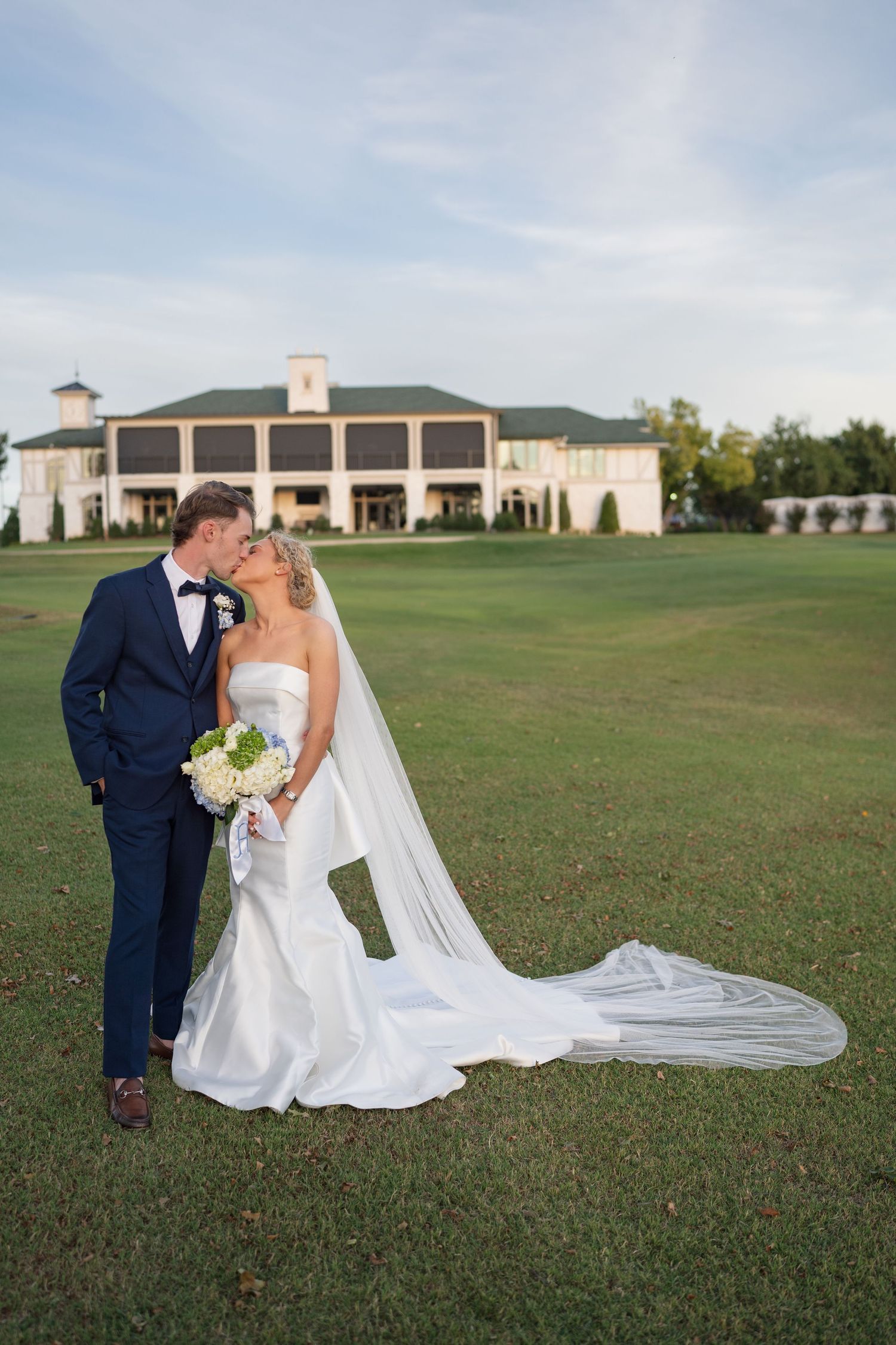 A wedding couple poses on a golf course with a stately clubhouse in the background.