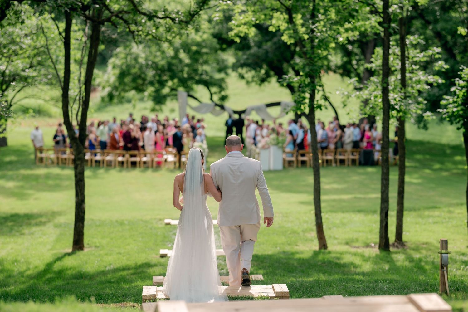 Bride and groom walk hand-in-hand toward outdoor wedding ceremony guests seated among lush green trees.