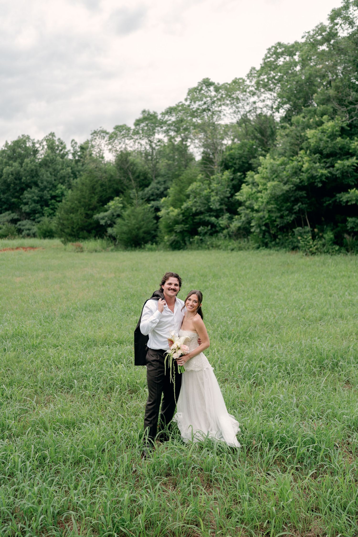 Bride and groom posing together in lush green meadow, bride in flowing white gown holding bouquet.