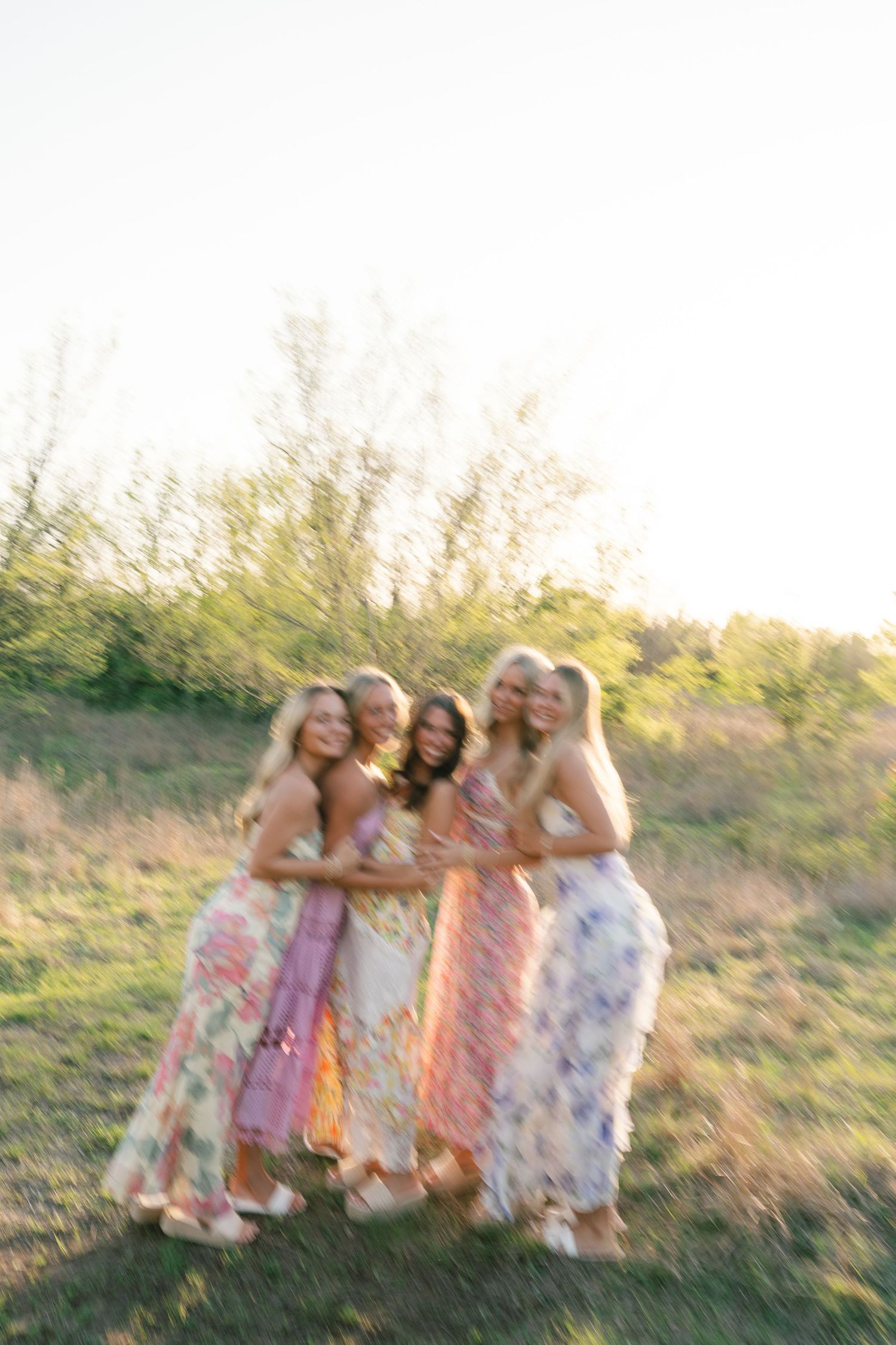 Group of friends in floral dresses embrace while laughing in sunlit field.