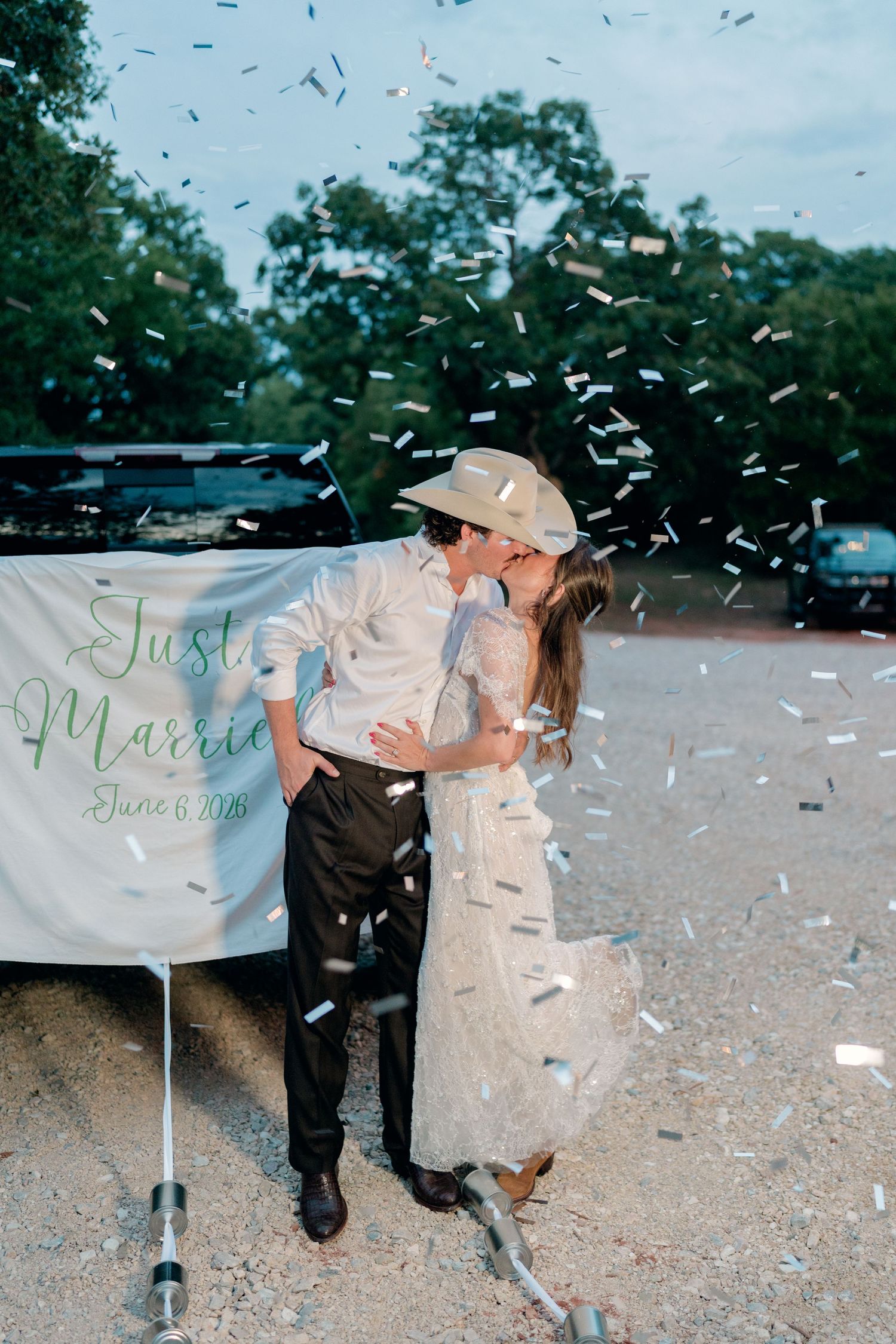 Newlyweds kissing under confetti shower beside Just Married banner at outdoor wedding celebration.
