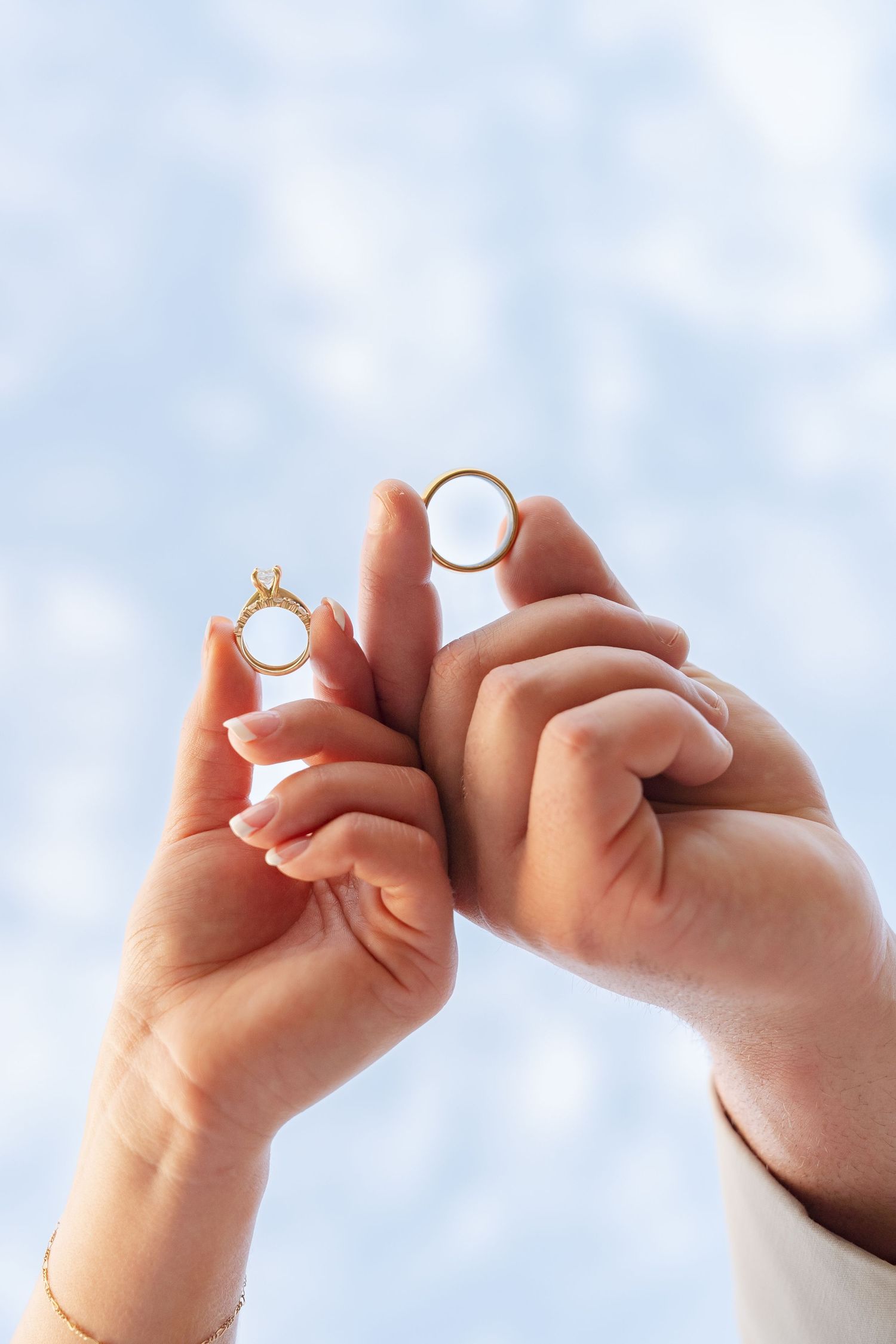 Hands holding wedding rings against a bright blue sky background.