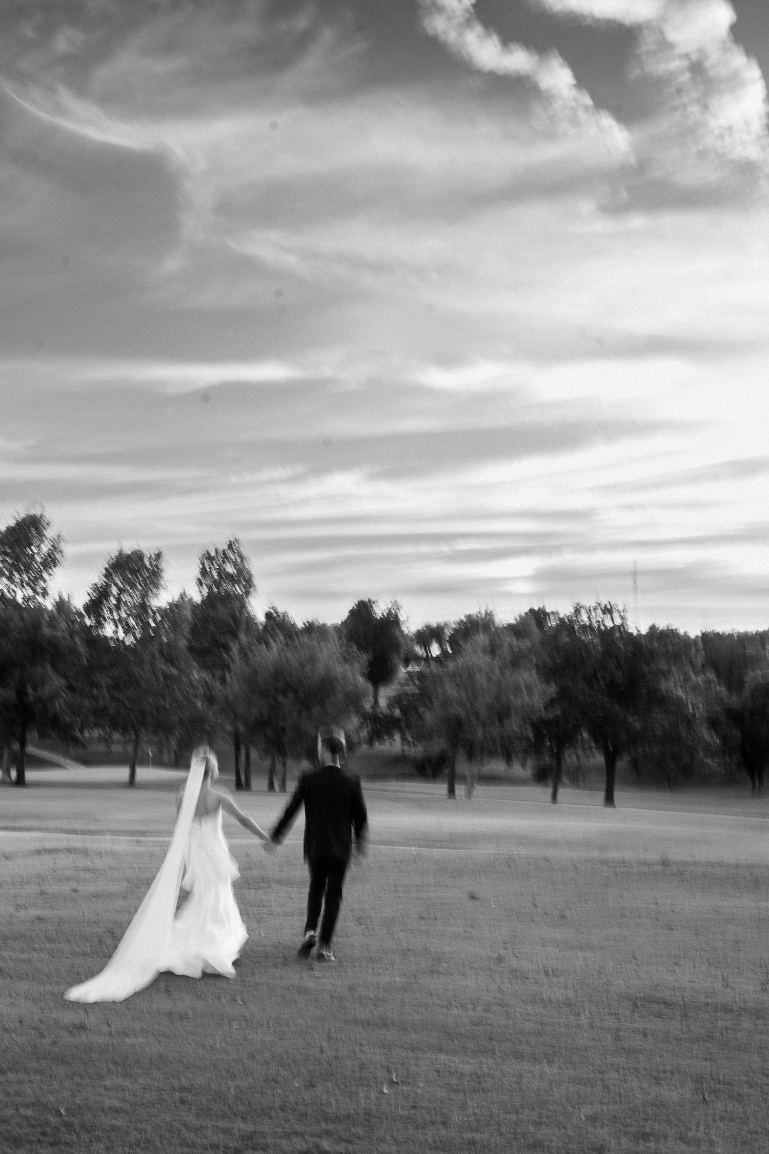 Black and white photo of newlyweds walking across a golf course under dramatic clouds.