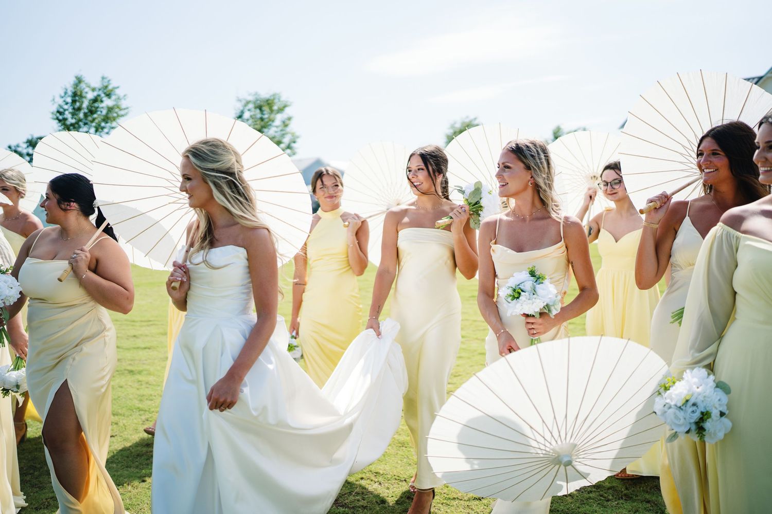 Wedding party poses with white parasols in a sunny outdoor setting.