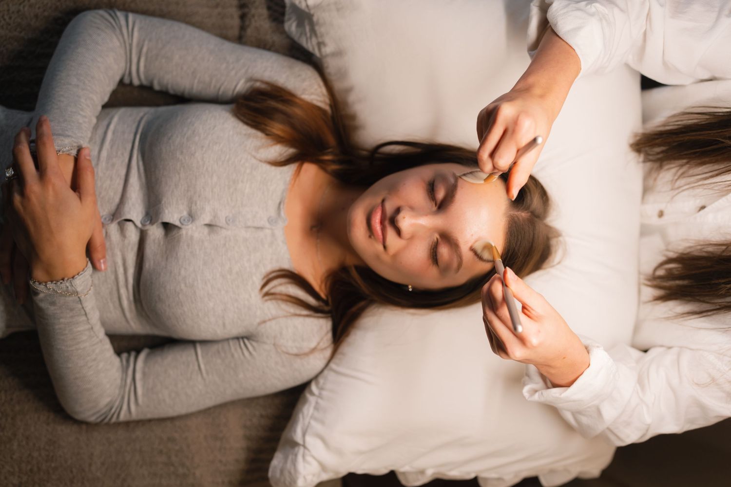 Woman with long dark hair lies on white bedding in various relaxed poses wearing a cozy gray sweater.