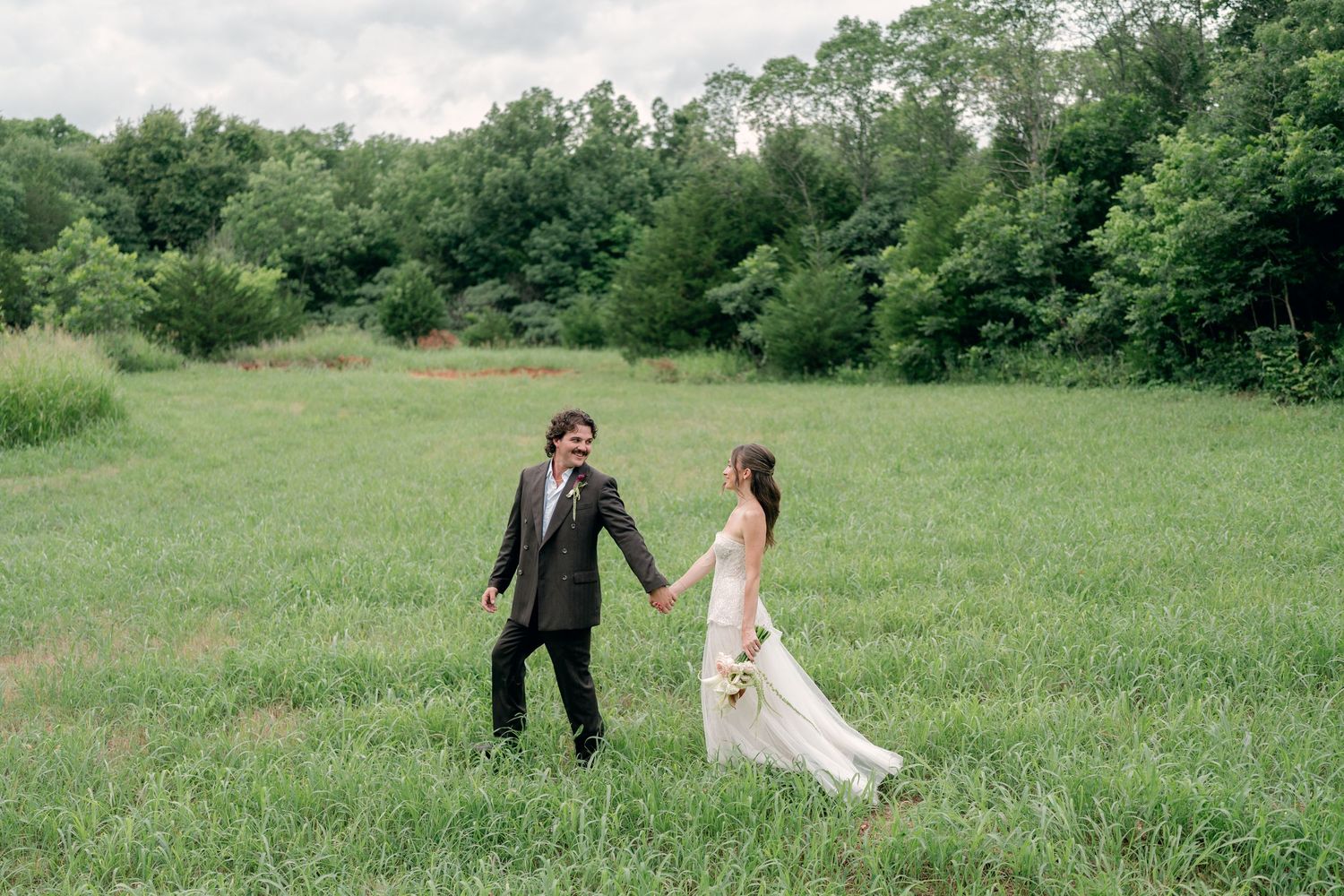A bride and groom hold hands walking through a lush green meadow with trees in the background on their wedding day.
