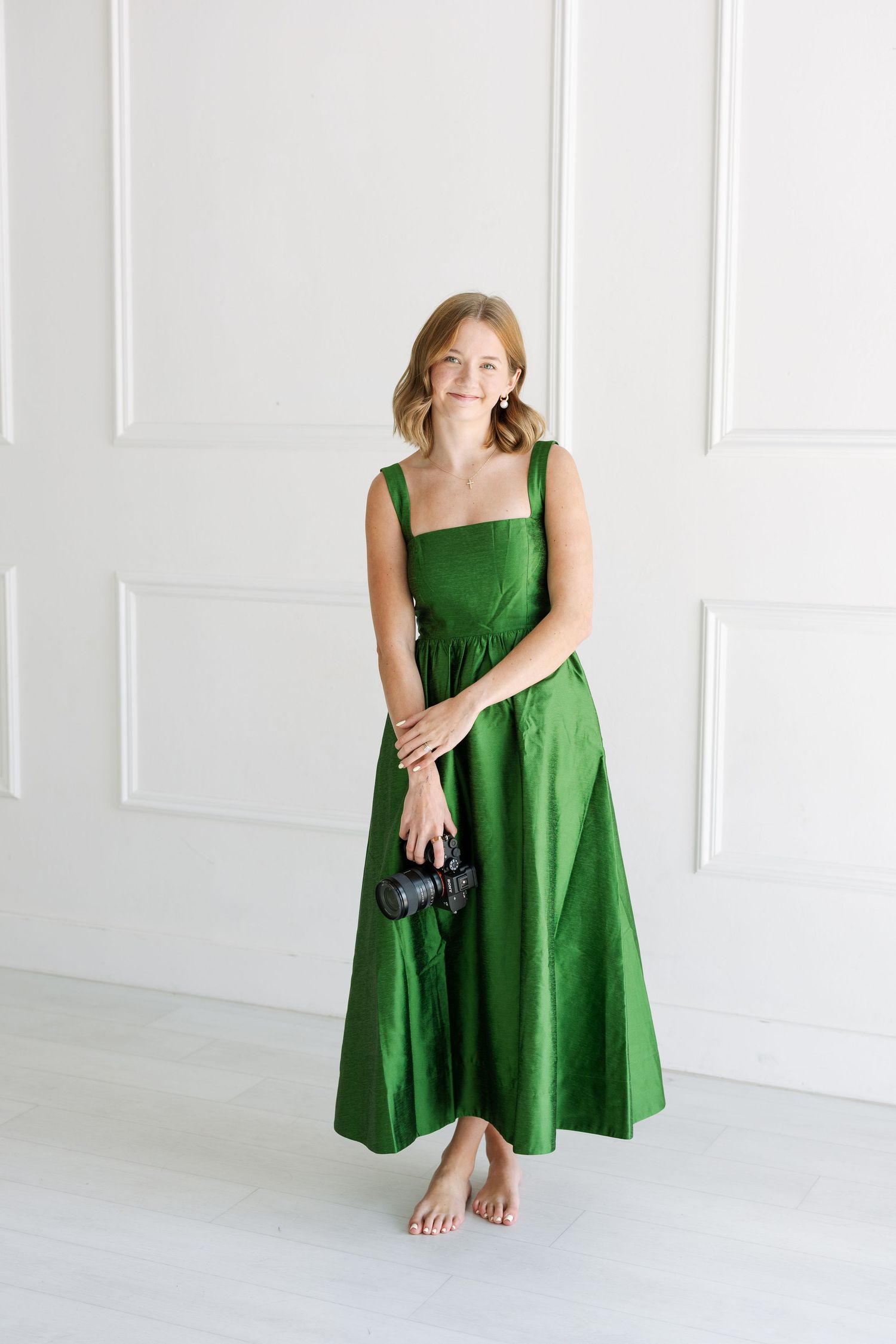 Woman in elegant green midi dress holding camera, standing barefoot against white wall in bright studio setting.
