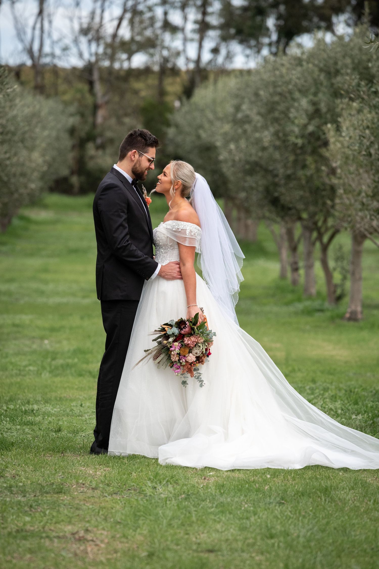 Romantic wedding portrait of couple standing among olive trees in an outdoor garden setting.