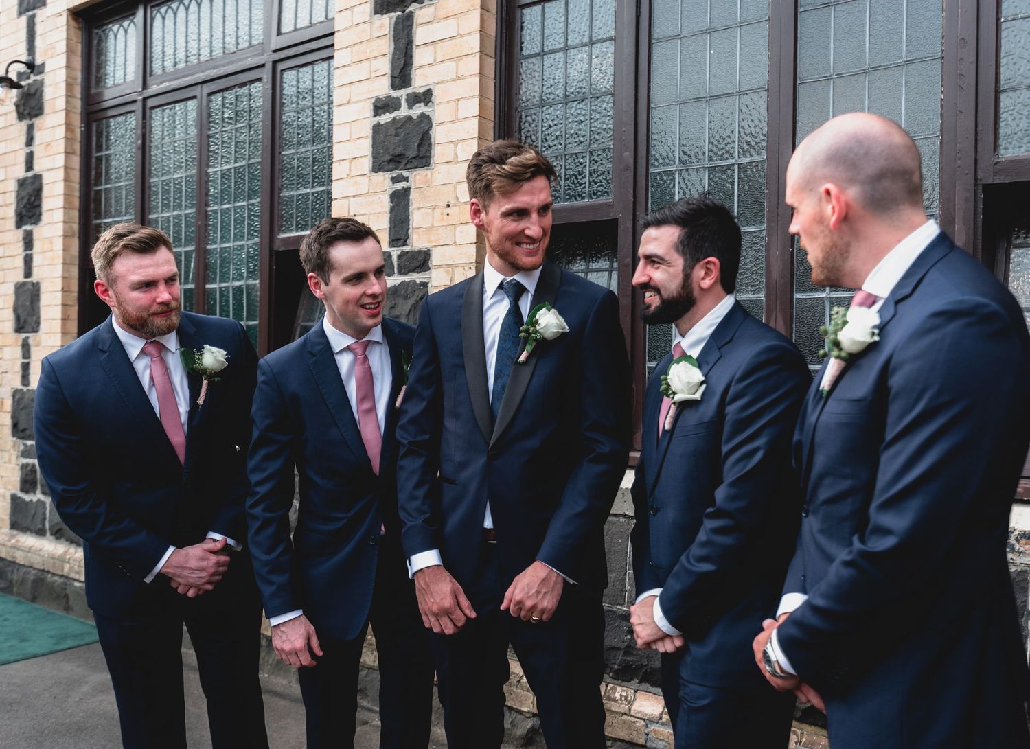 Groomsmen in black suits stand in a row outside a building with large windows while sharing laughs together.