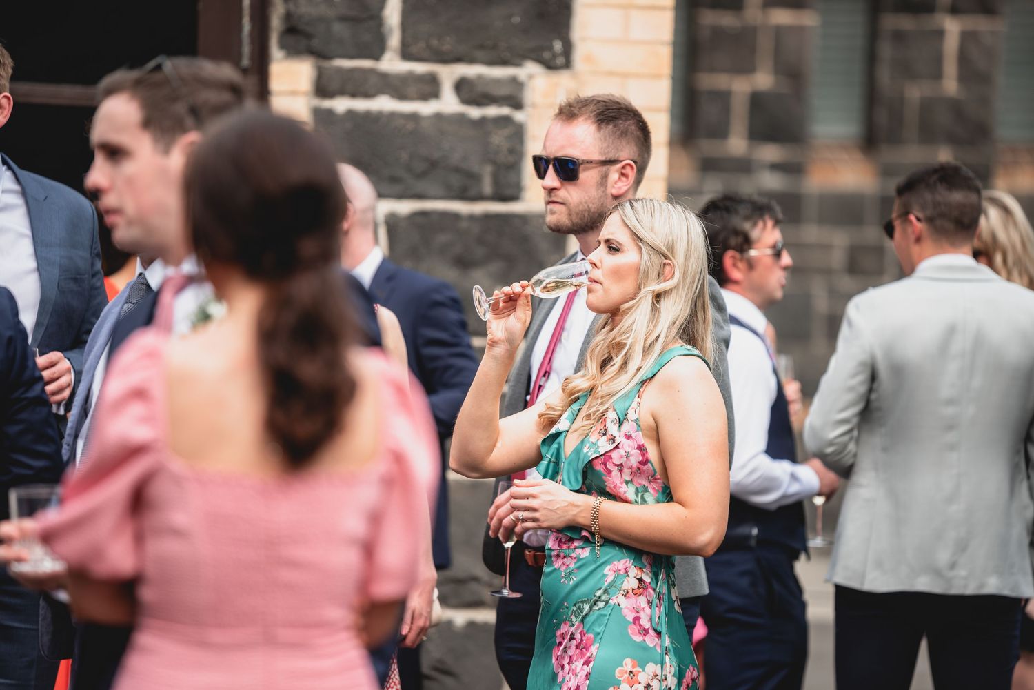 A group of wedding guests mingling and socializing at an outdoor reception with stone walls in the background.