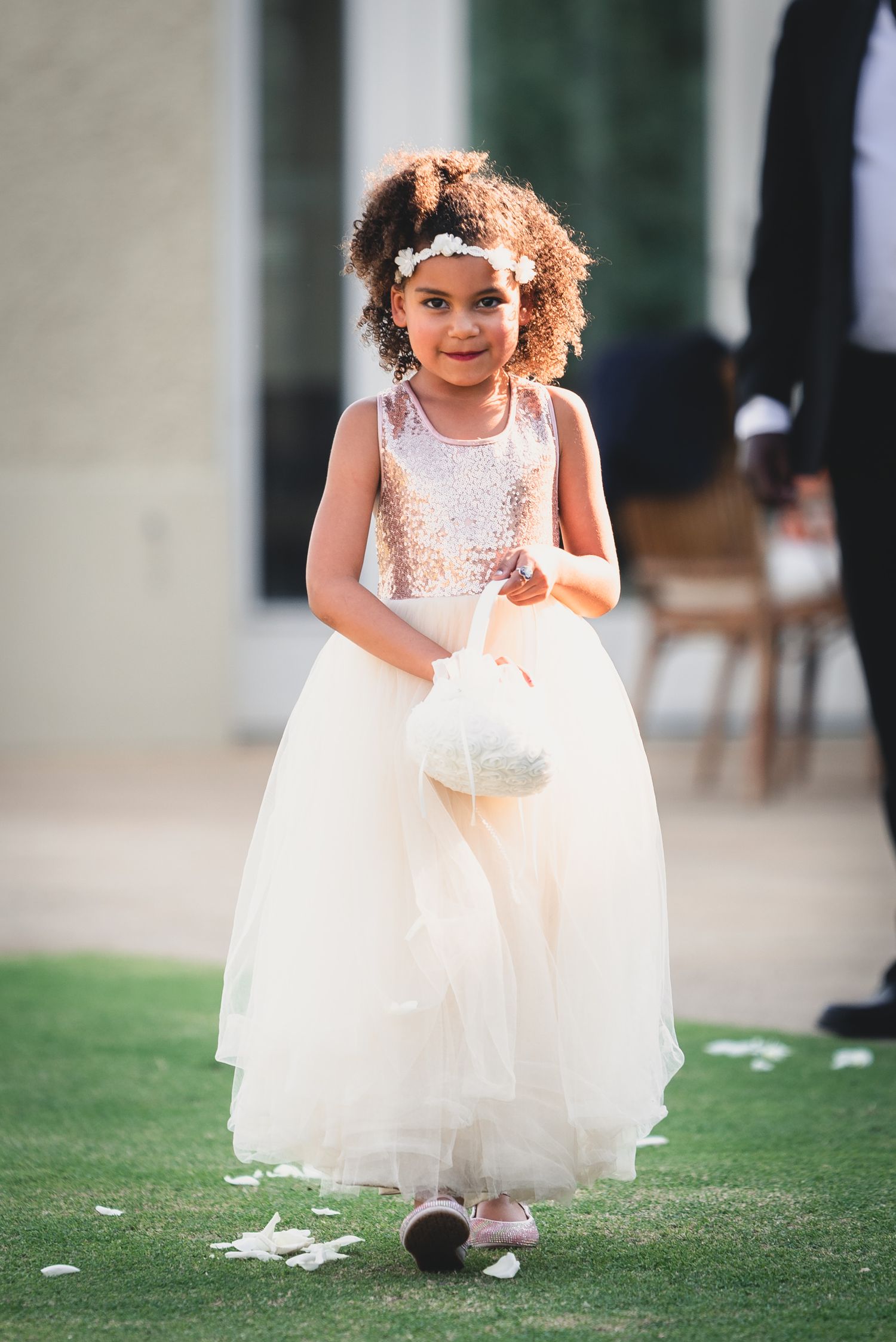 A young flower girl in a white dress and curly hair skips playfully across a grassy lawn at an outdoor wedding.
