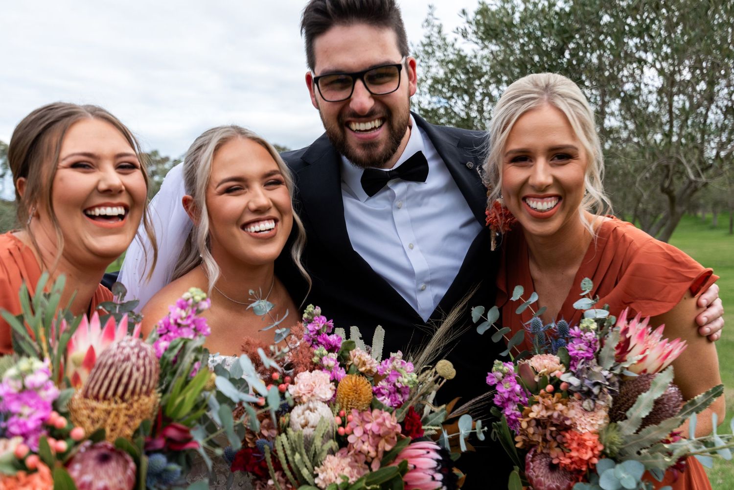 Wedding party members smile and laugh together while holding vibrant pink and coral floral bouquets outdoors.