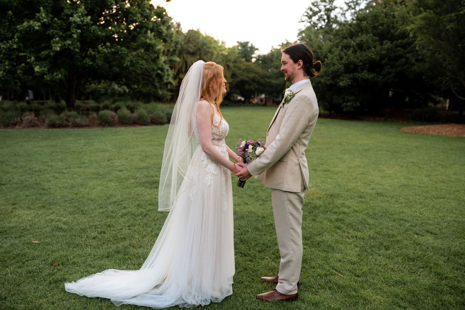 A romantic outdoor wedding ceremony with a couple in white dress and beige suit standing together on a green lawn.