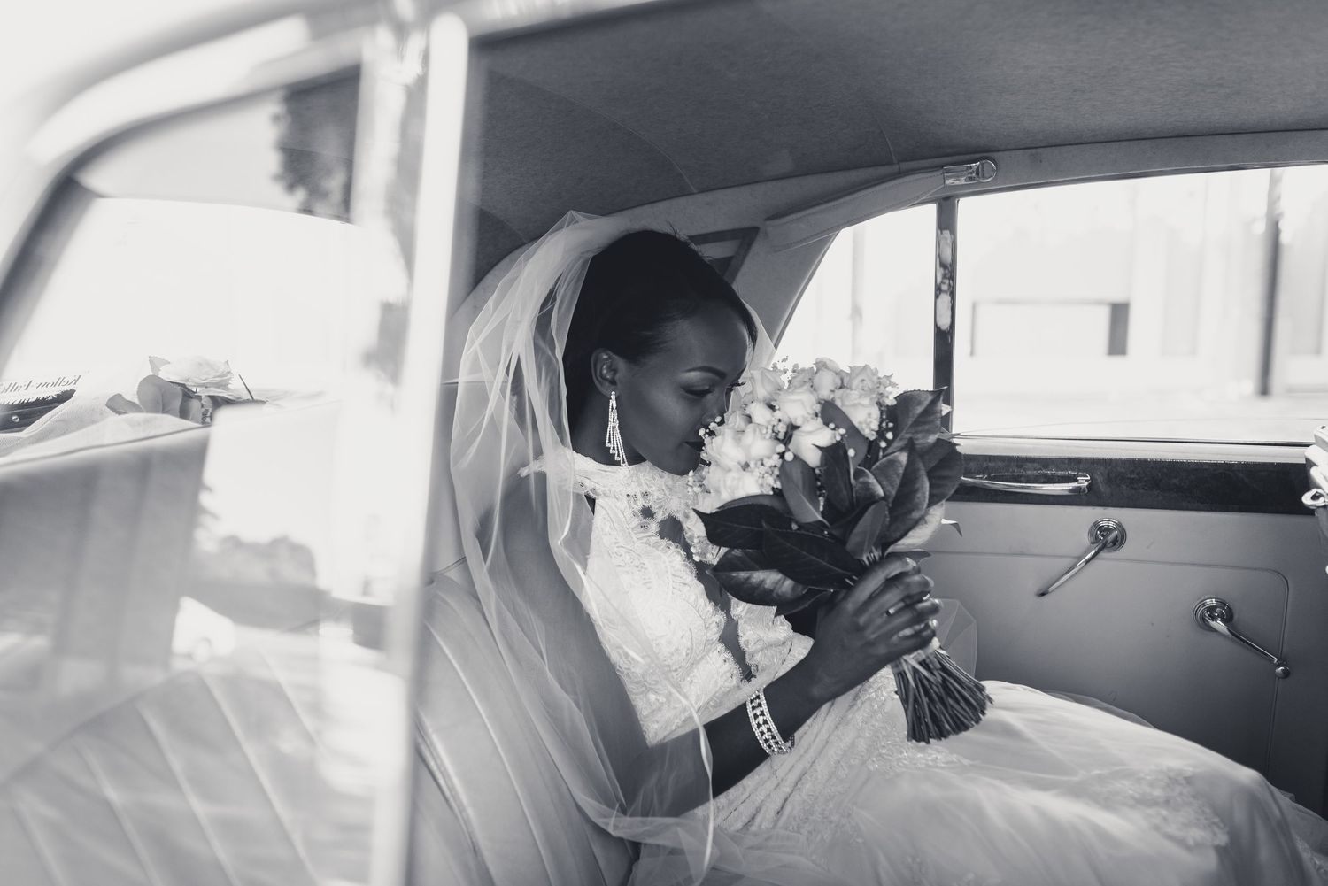 A bride in a white wedding dress and veil sits in a vintage car while holding a bouquet of flowers.