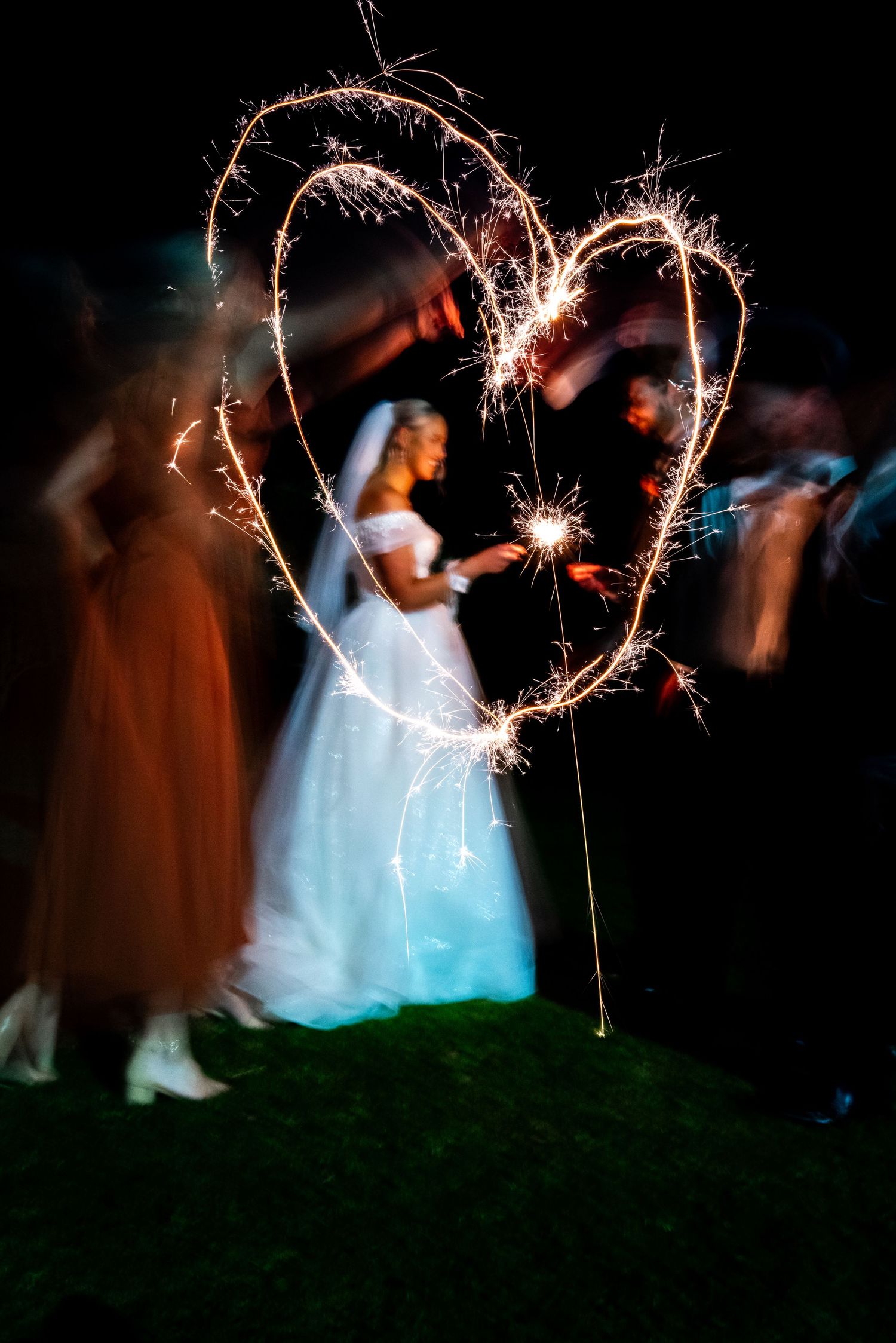 A sparkler creates a heart shape light trail against a dark background at a wedding celebration.