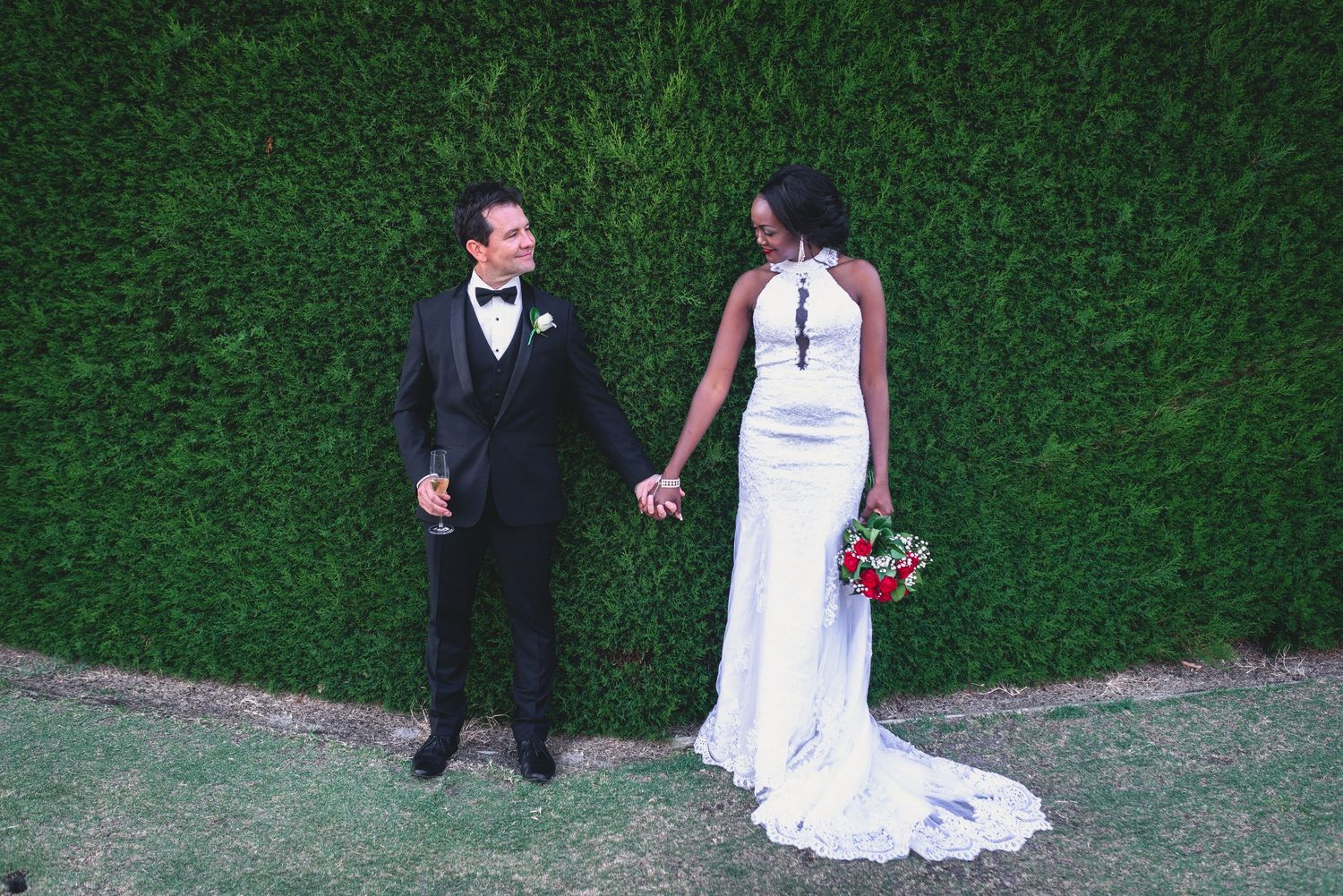 A couple poses for wedding photos in front of a manicured green hedge wall at a garden venue.
