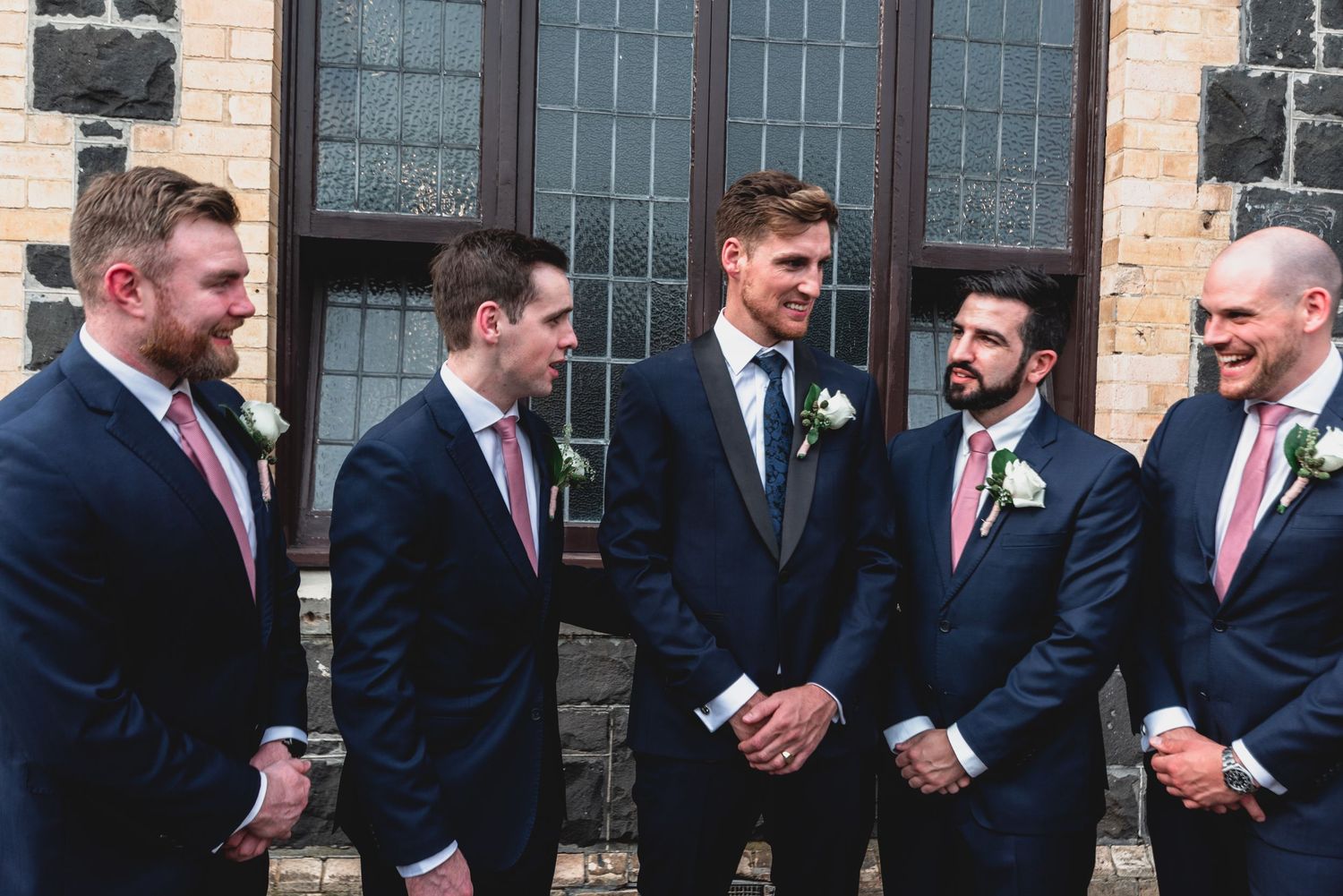 A group of groomsmen in navy suits and pink ties share a candid moment outside a brick building.