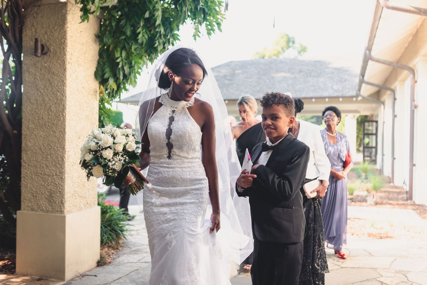 A wedding party walks down a covered outdoor walkway with stone columns and greenery overhead.