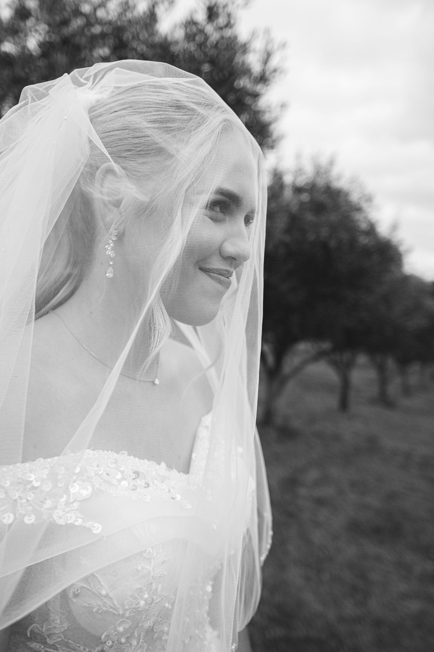 A bride wearing a delicate white veil poses outdoors in a romantic black and white wedding portrait series.
