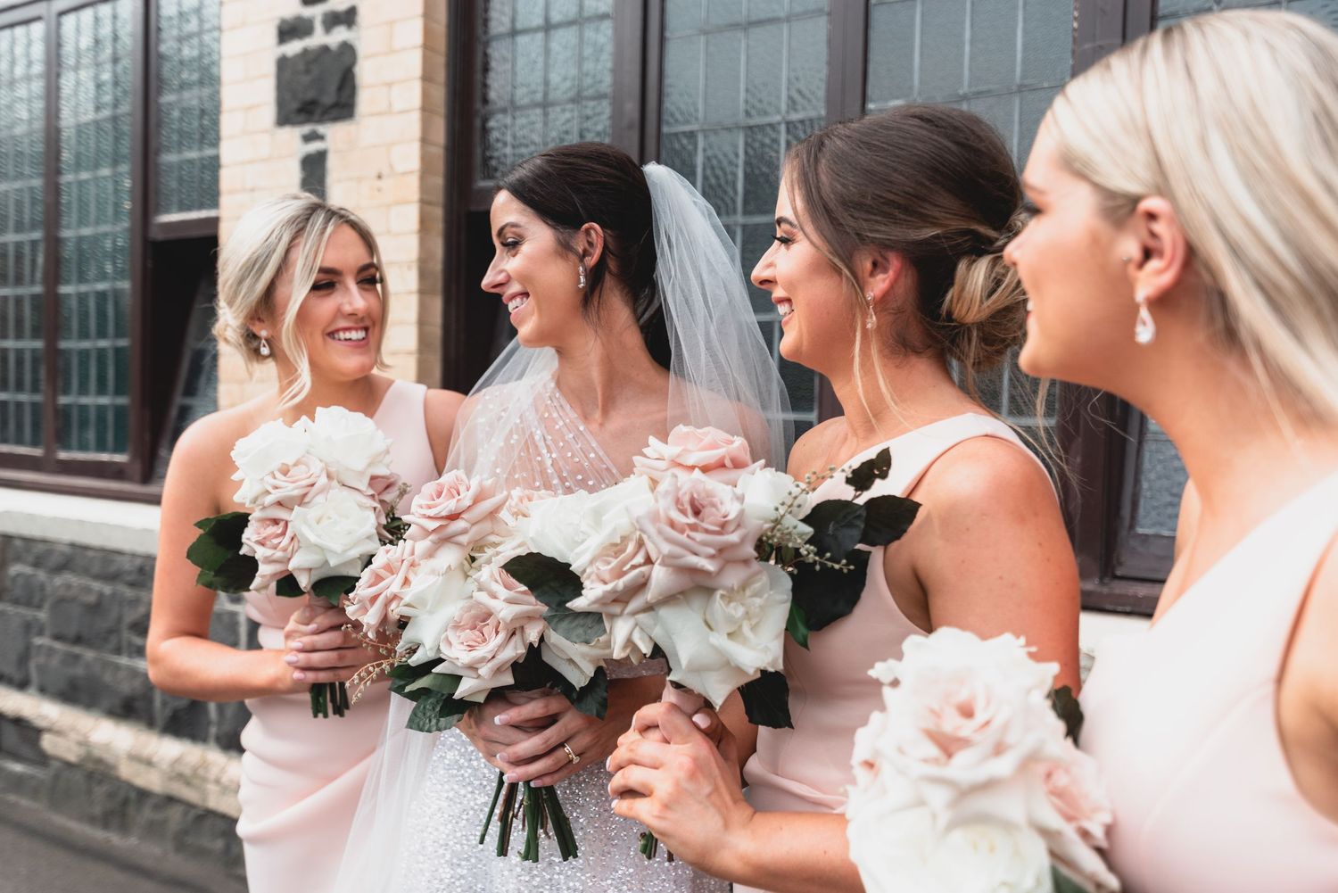 Bridesmaids in light pink dresses holding elegant rose bouquets smile together outdoors.