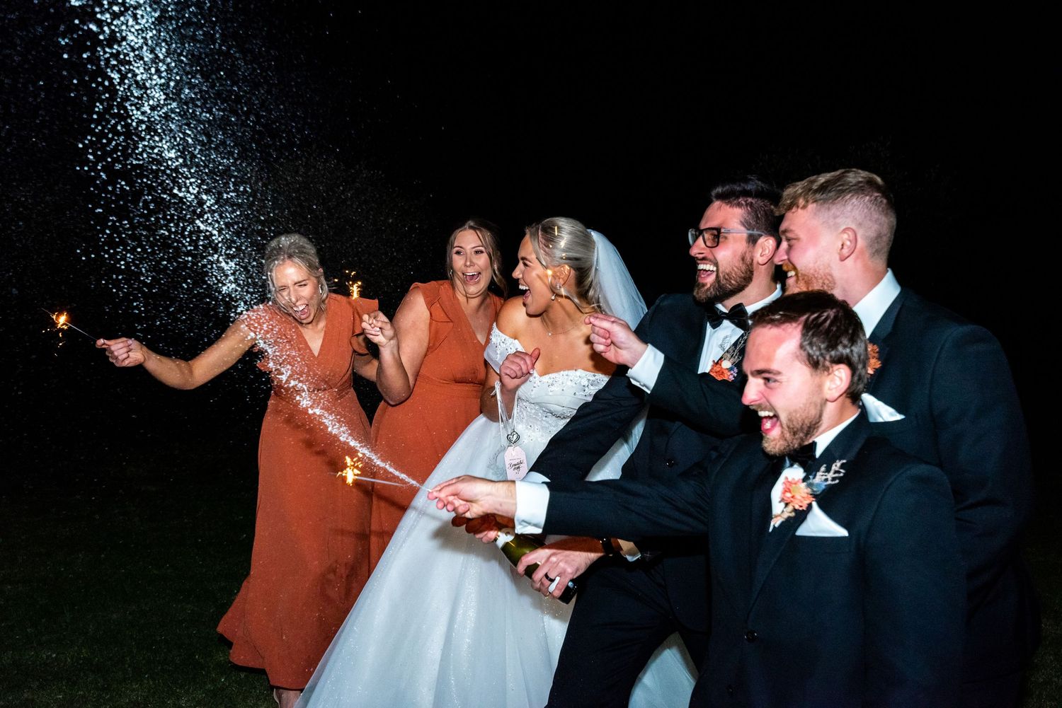 Wedding party laughs while holding sparklers during outdoor nighttime celebration.