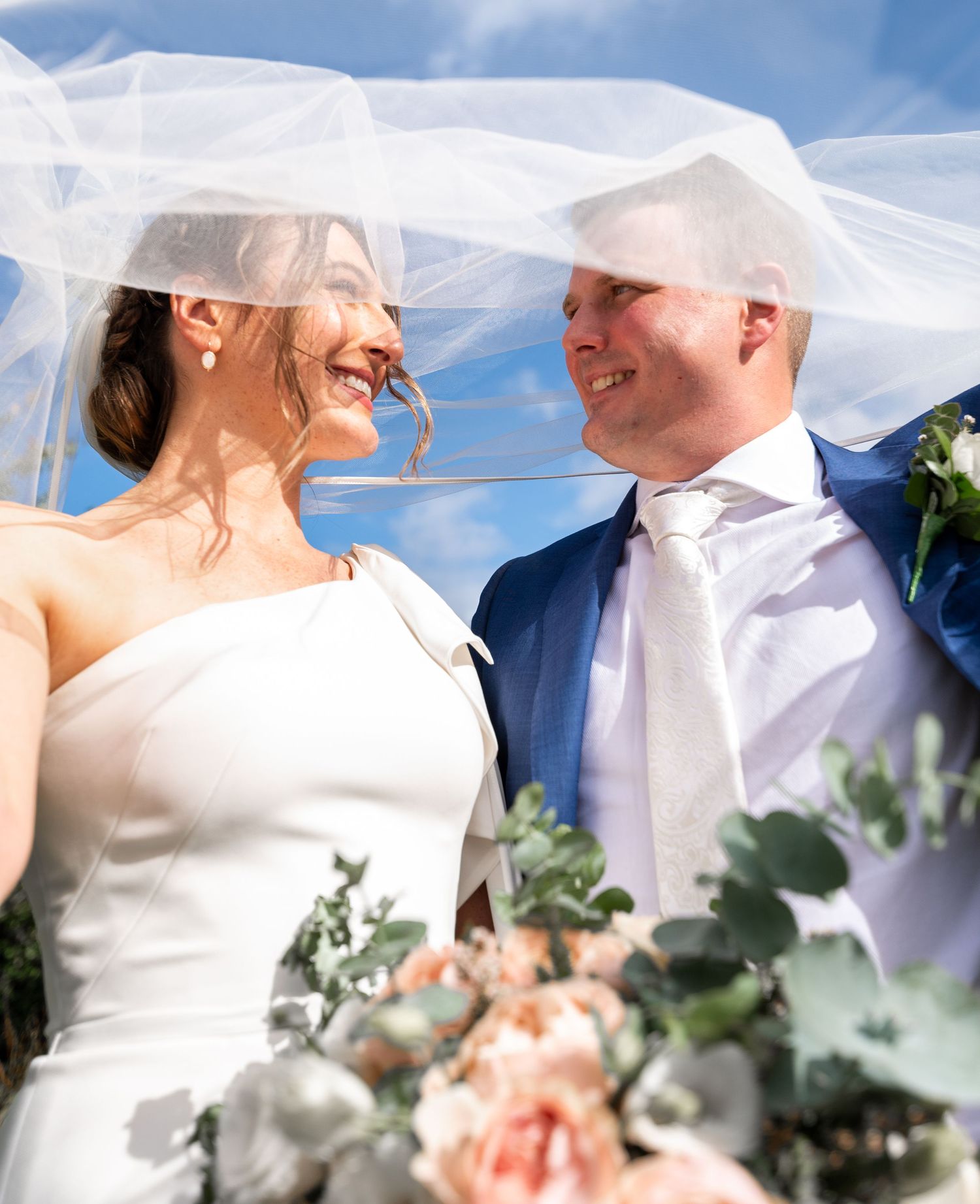 A romantic wedding couple shares intimate moments under a flowing white veil with soft pink florals and greenery.
