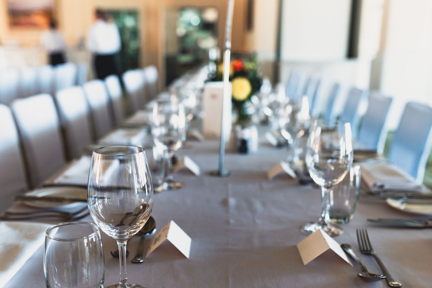 Elegant banquet table settings with white linens, wine glasses, and place cards in a modern restaurant dining room.