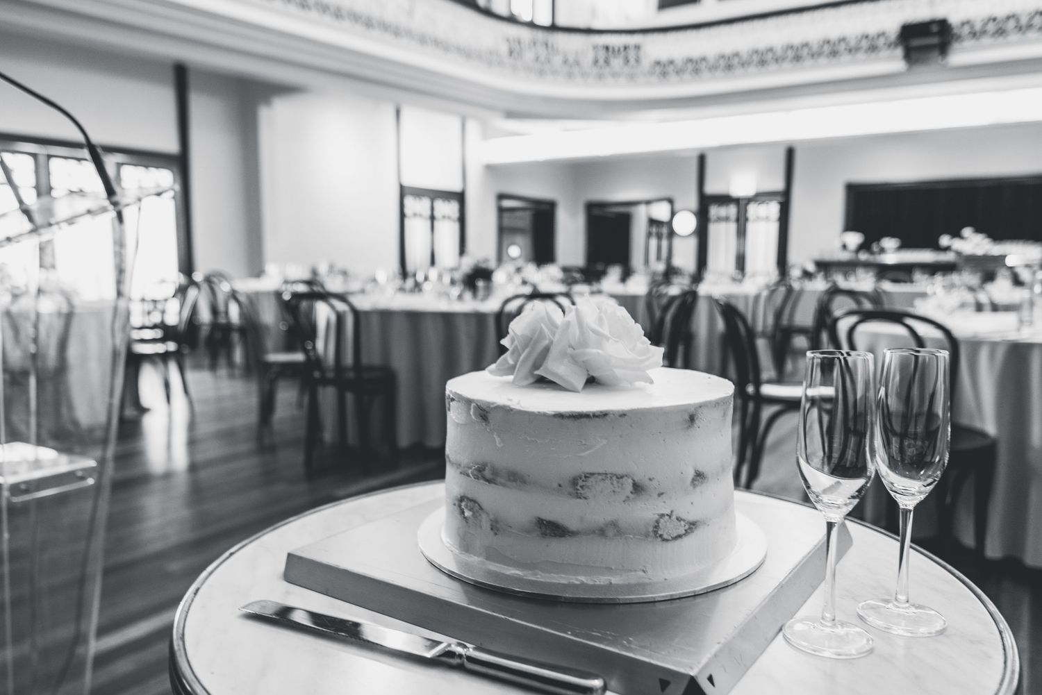 Black and white photo of an elegant wedding cake displayed on a table with champagne glasses in a formal dining room.
