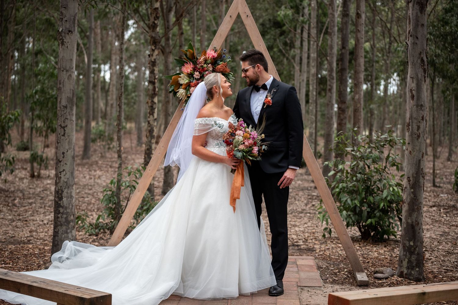 Bridal party poses in a wooded forest setting with rustic wooden arch and natural decor.