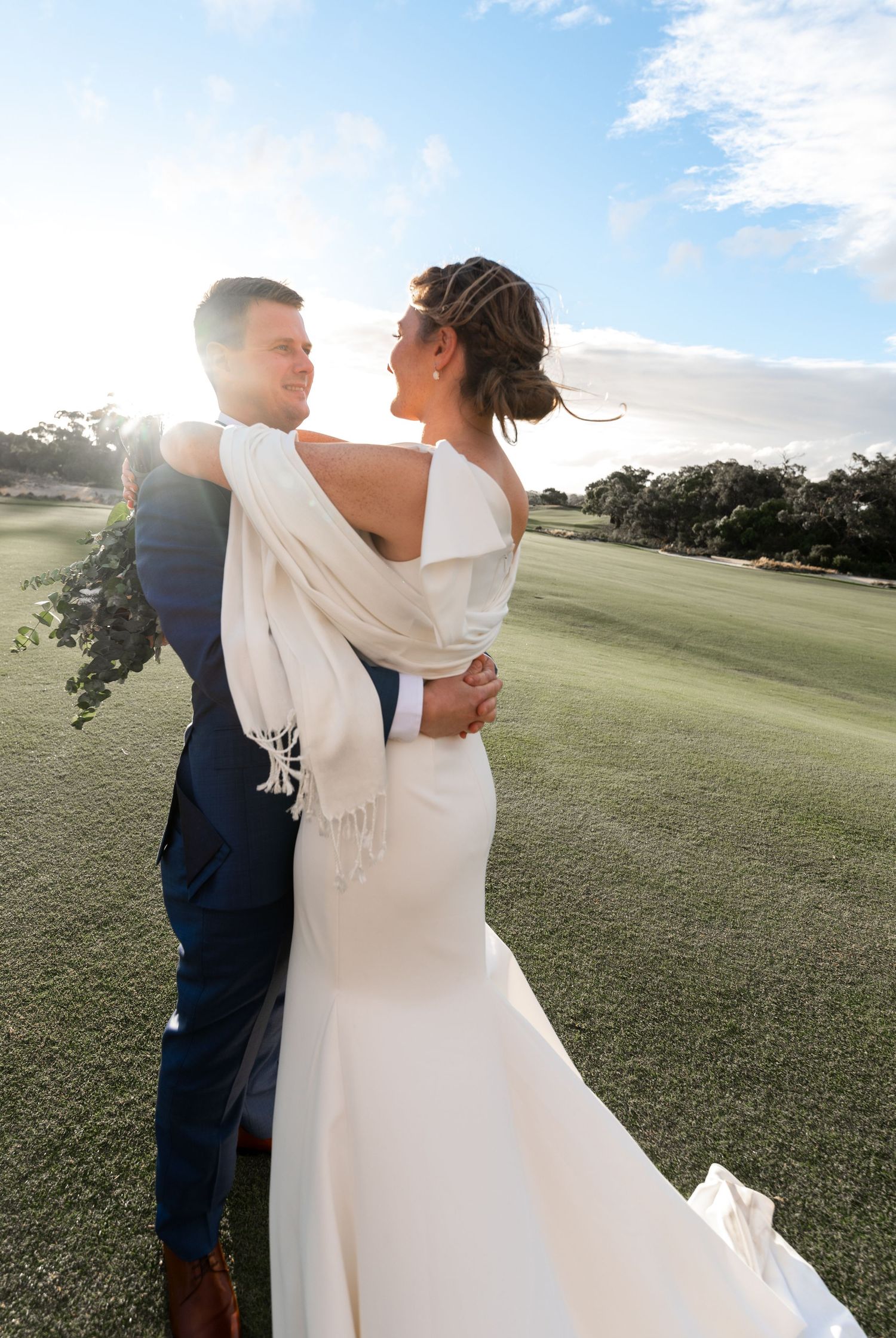 A newlywed couple embraces in a sun-drenched field during their outdoor wedding photoshoot.