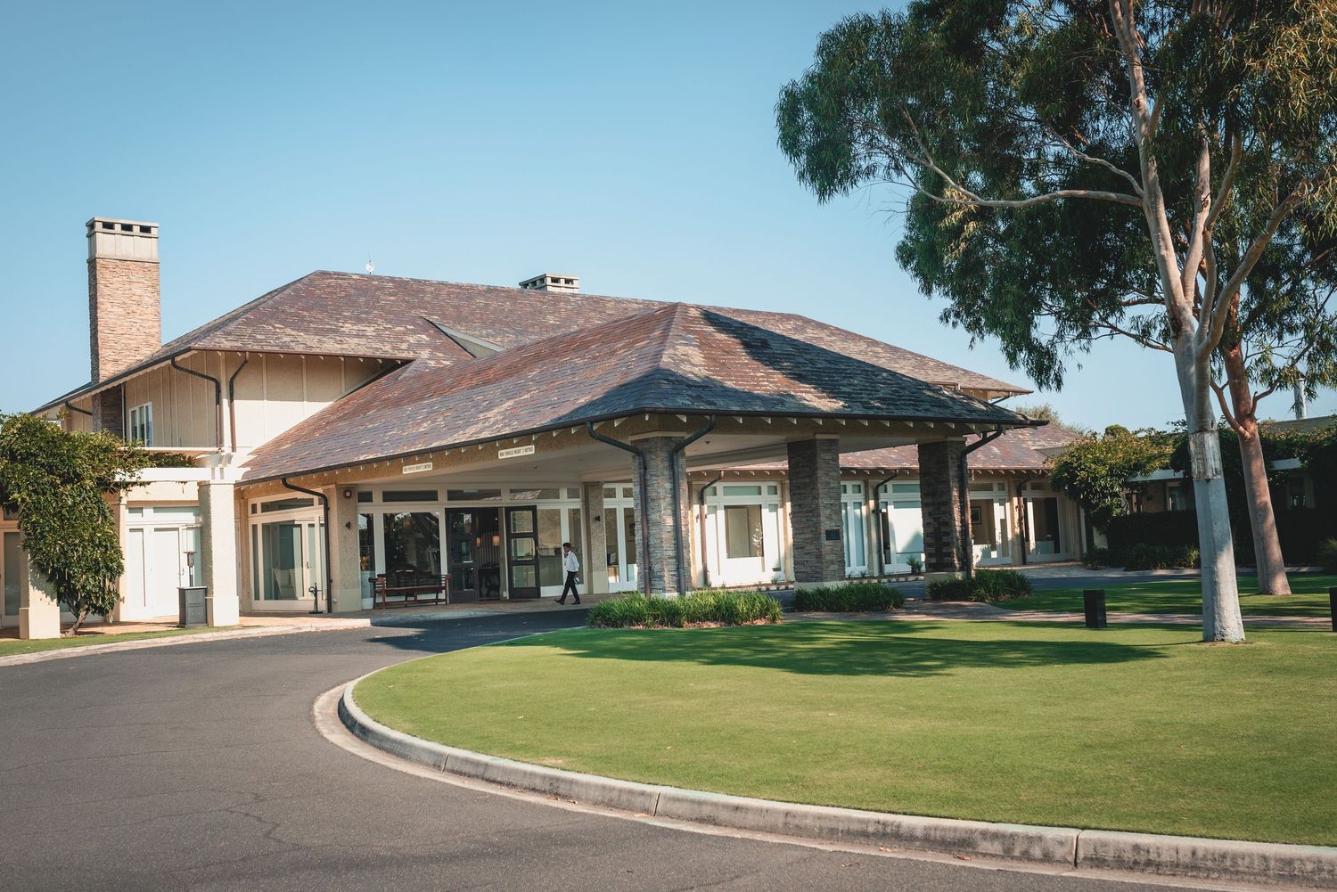 A curved driveway leads to an elegant beige clubhouse with a covered entrance and manicured lawn.