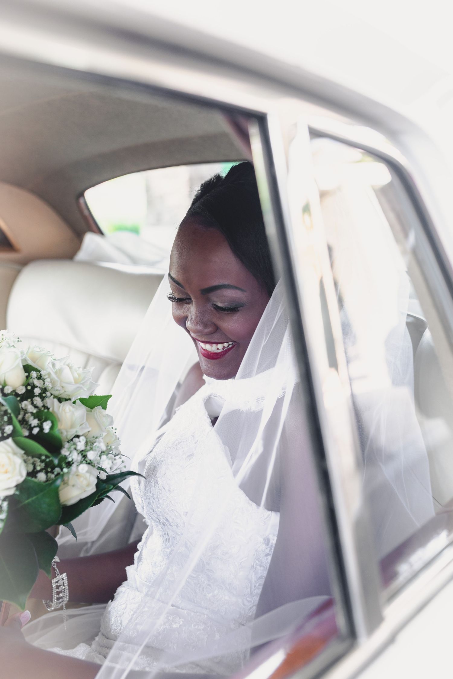 A bride in a white wedding dress and veil smiles while sitting in the back seat of a car.