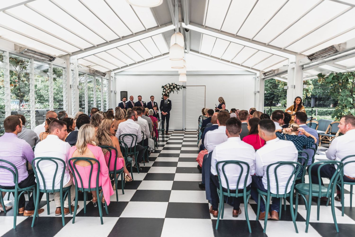Panoramic view of guests seated in rows at an outdoor wedding ceremony under a white canopy structure.