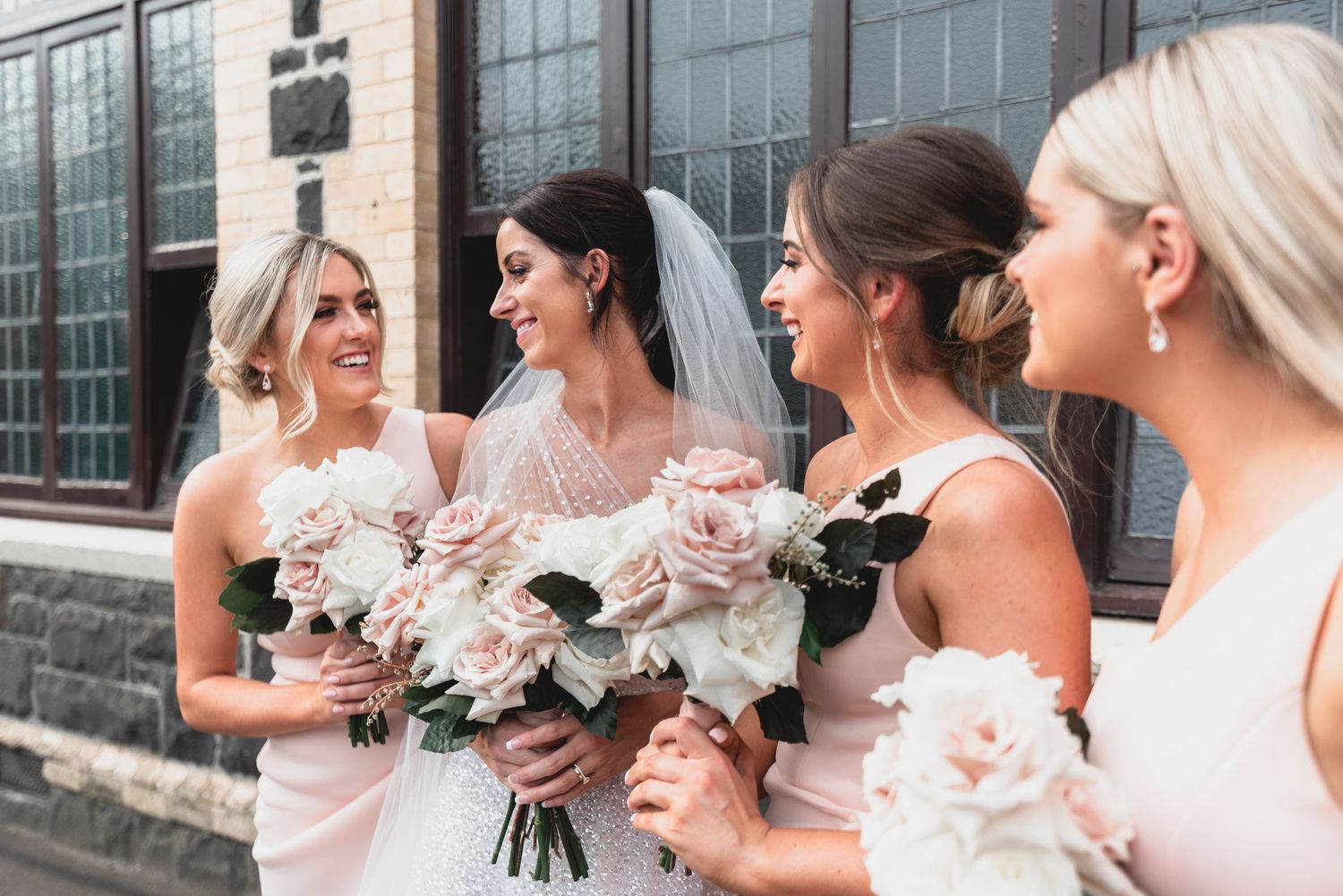 Bridesmaids in blush pink dresses laugh together while holding elegant white floral bouquets outside a building.