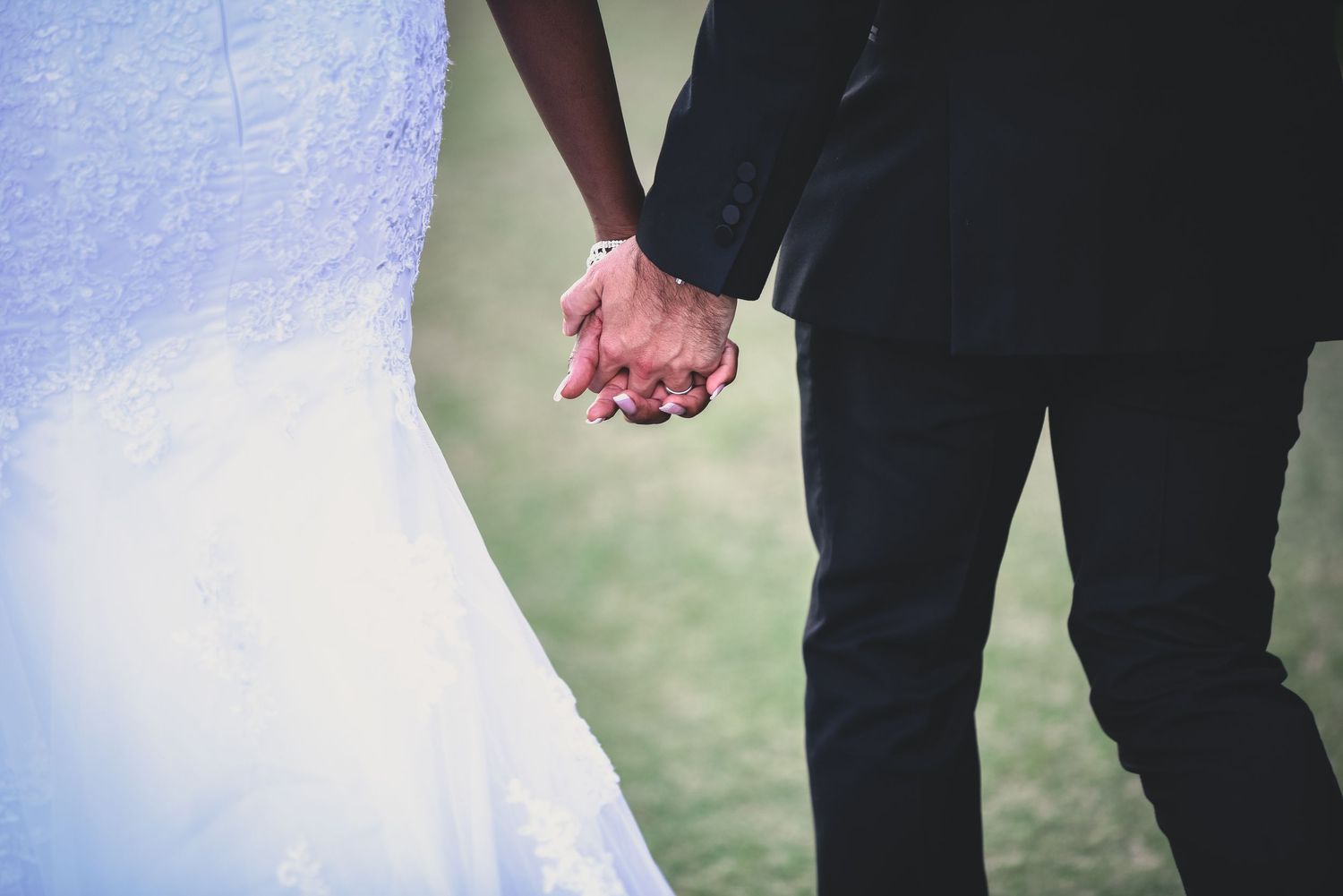 Couple holding hands in formal wedding attire standing on grass, showing close-up details of dress and suit.