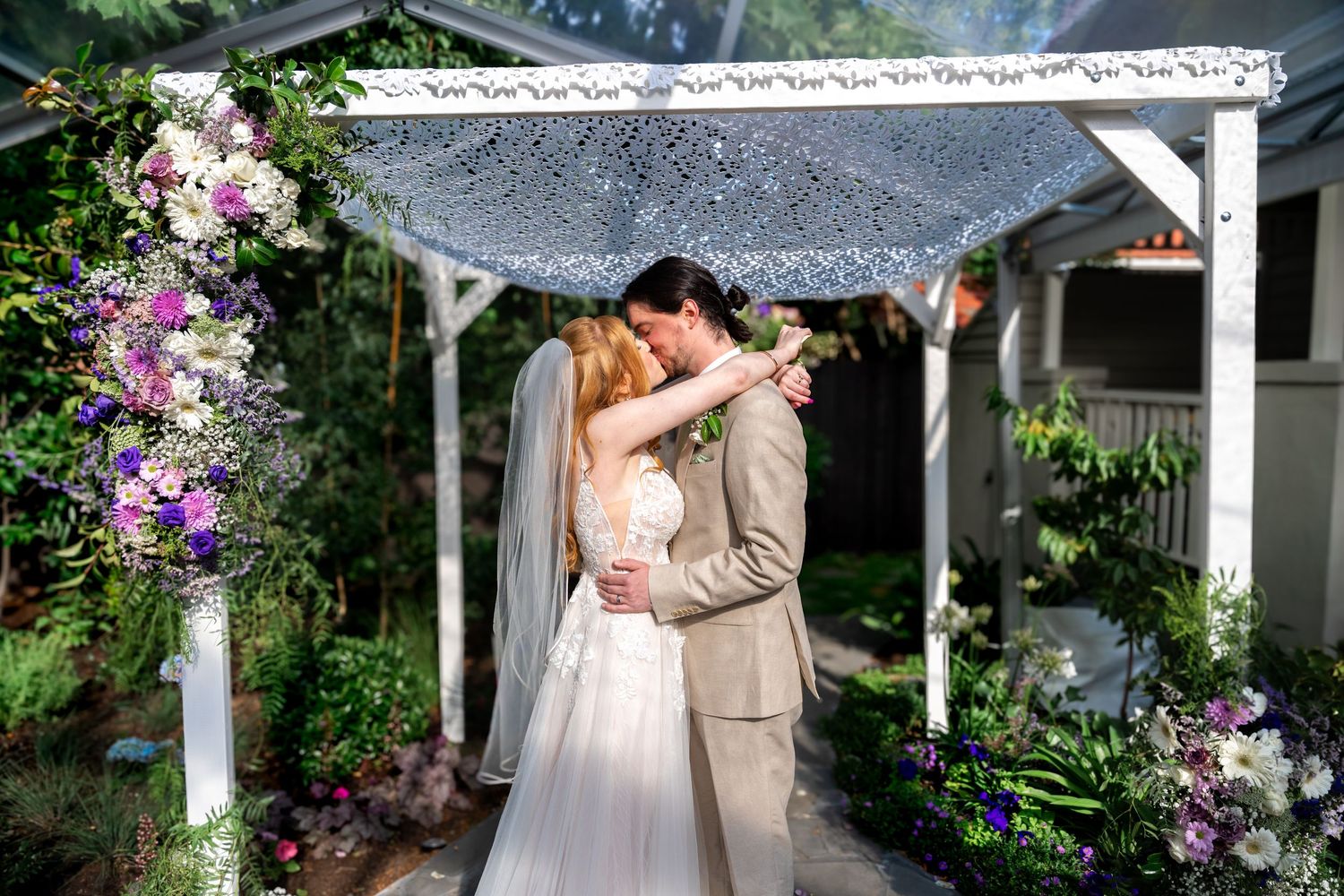 A romantic wedding couple embraces under a floral-covered garden pergola on their special day.