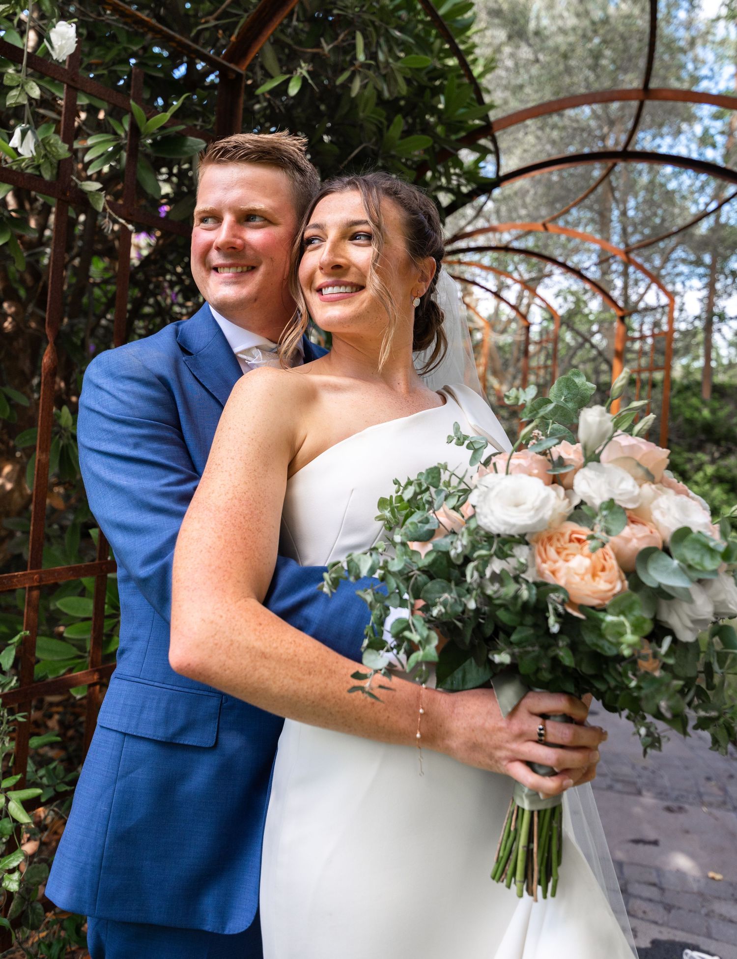 A wedding couple poses together in a garden setting with a white floral bouquet and blue suit.