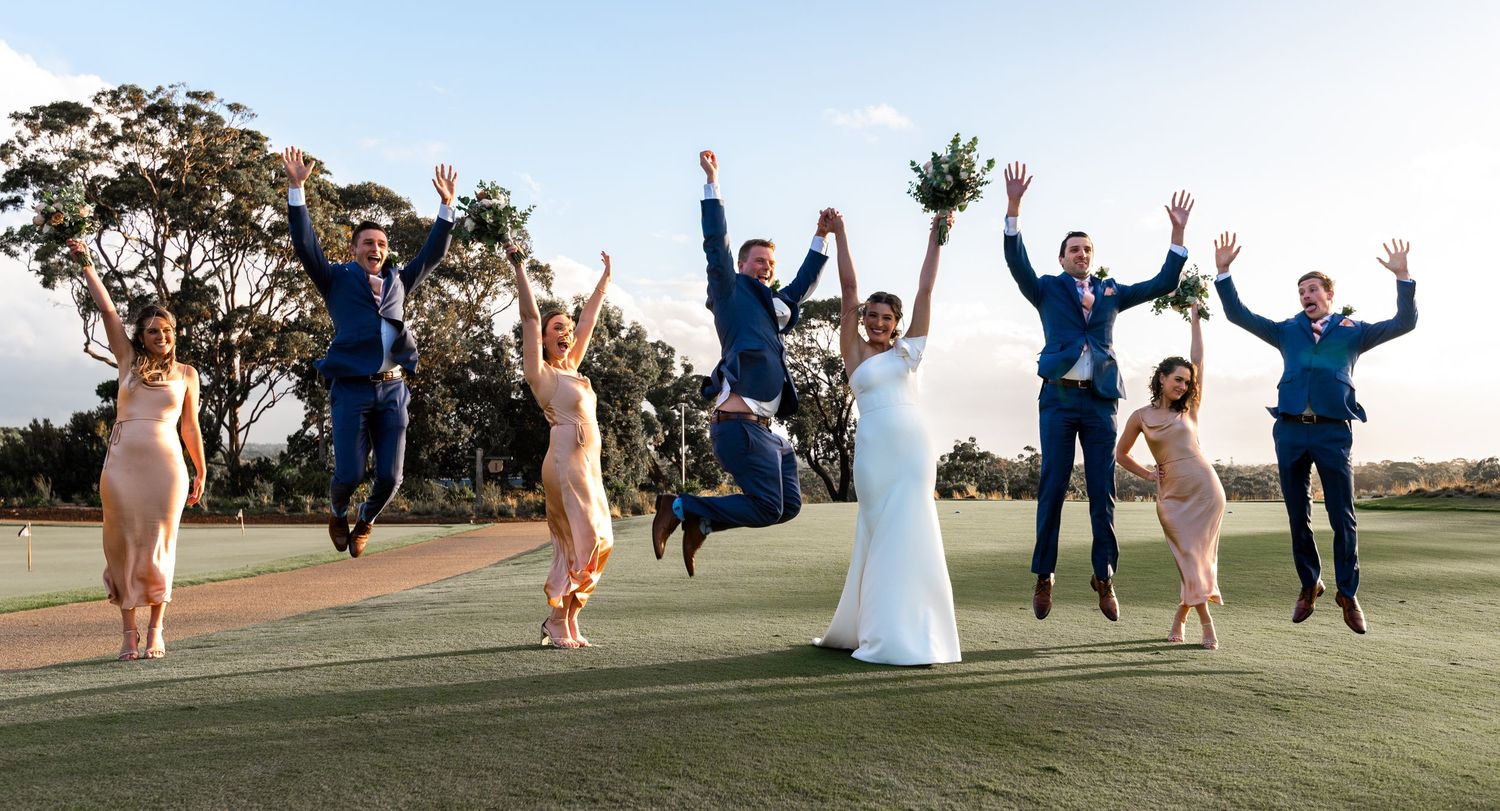 Wedding party members in navy blue attire jump joyfully together on a grassy hill at sunset.