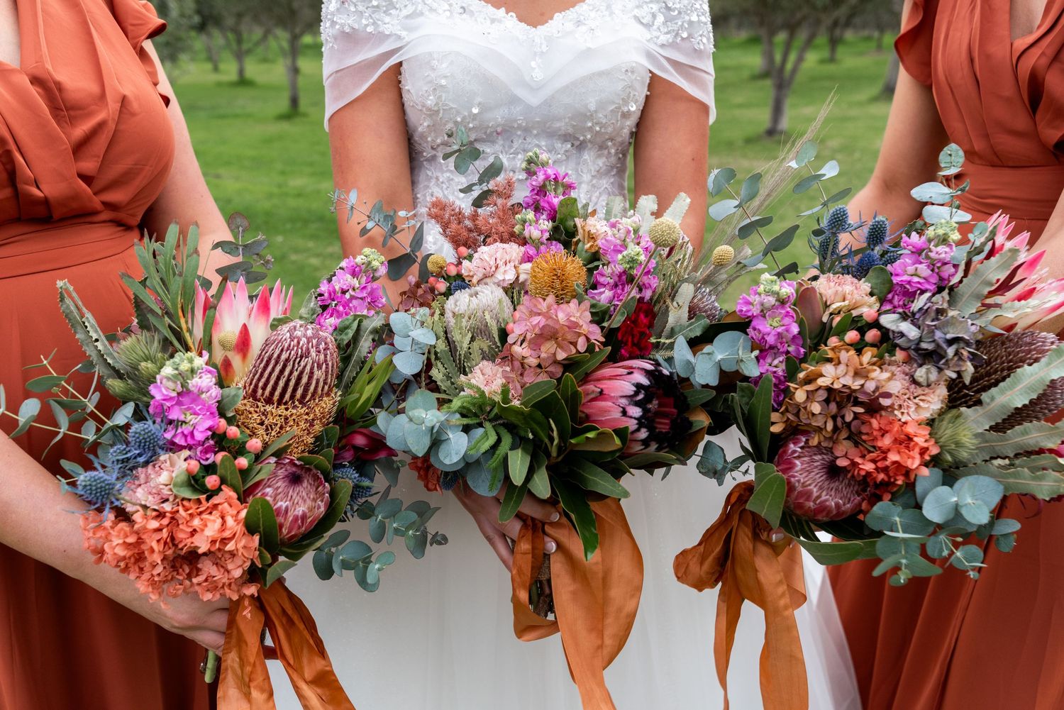 Close-up of vibrant wedding bouquets with pink, purple and coral flowers tied with orange ribbons against a white dress.