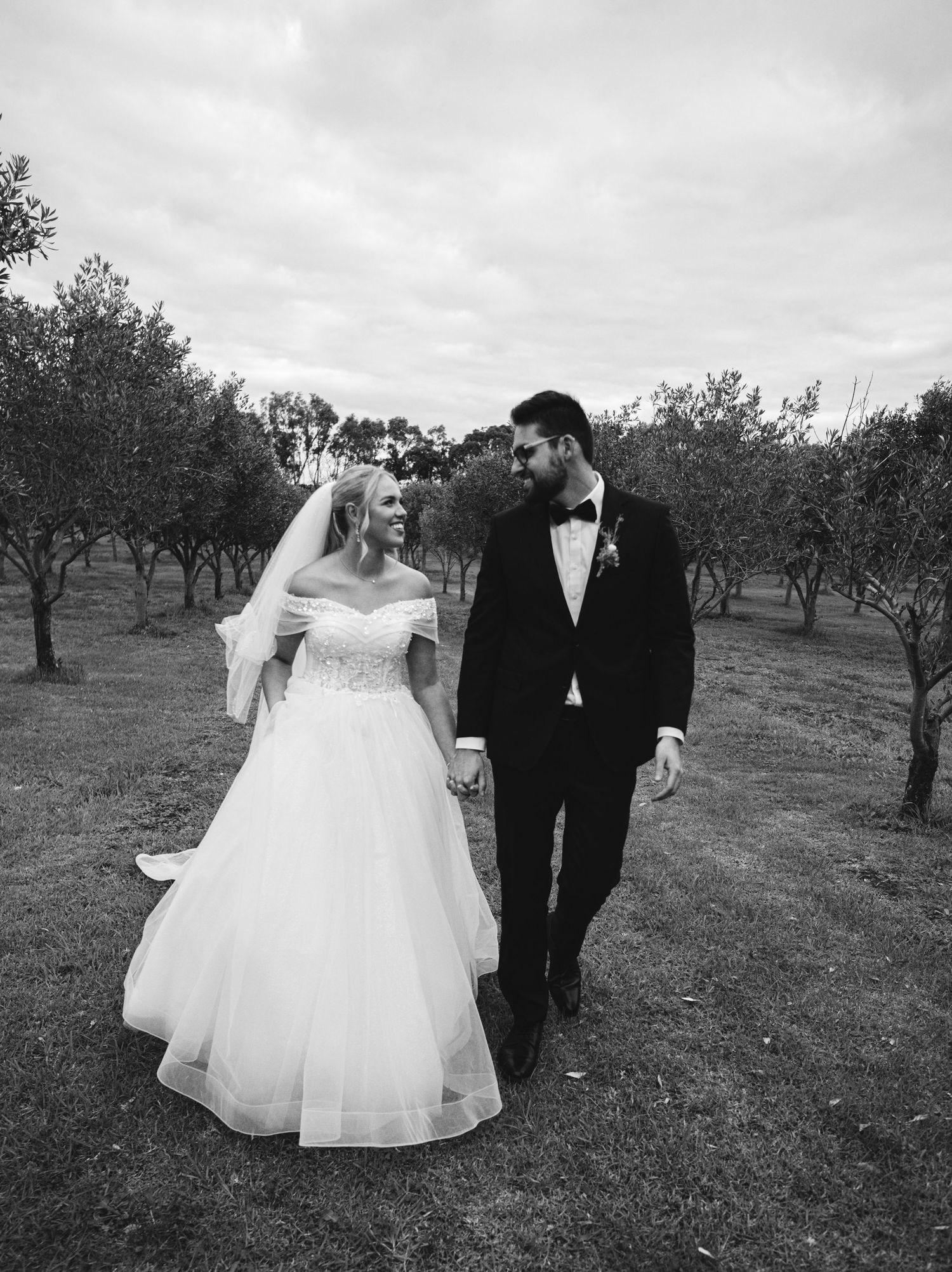 A couple in wedding attire walks together through an orchard on their special day in black and white.