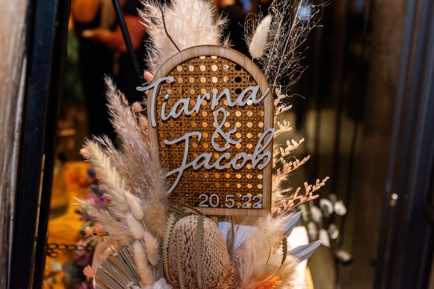 A decorative wedding sign with dried pampas grass and floral arrangements.