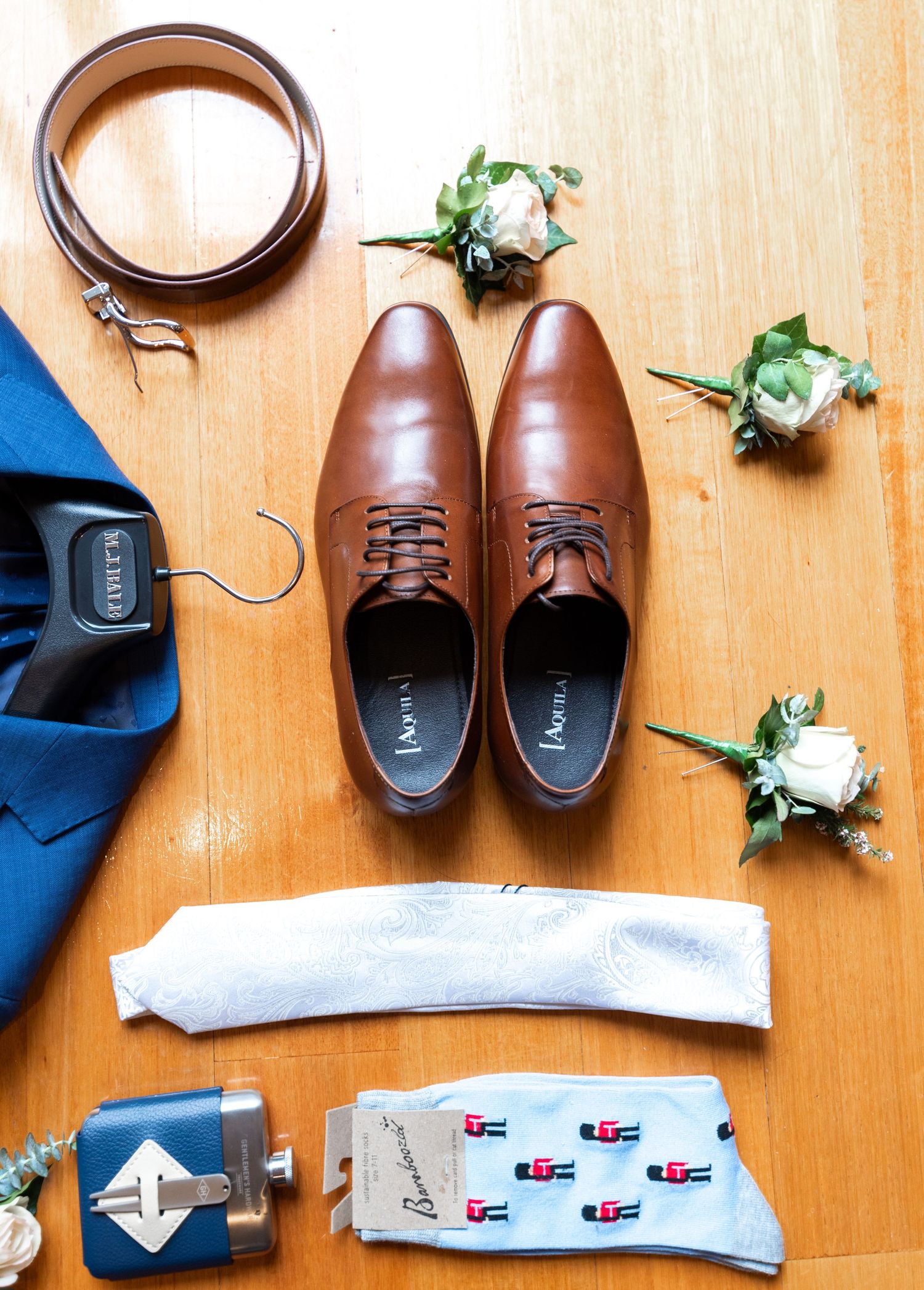 Brown leather dress shoes with matching belt, blue tie, socks and white boutonniere flowers arranged on wooden surface.