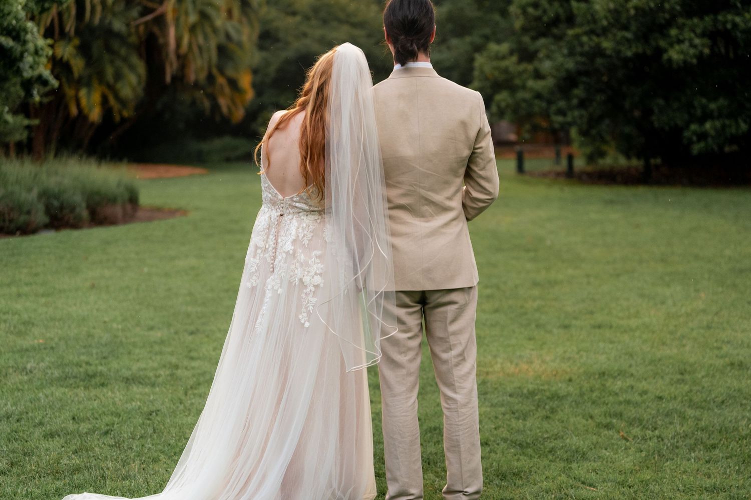 A couple stands together on a manicured lawn, with the bride's veil and dress flowing in the breeze.