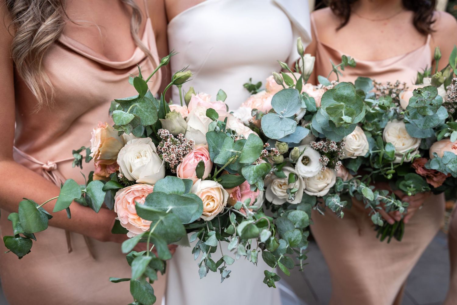 Wedding party members hold elegant bouquets featuring blush roses and cascading eucalyptus greenery.