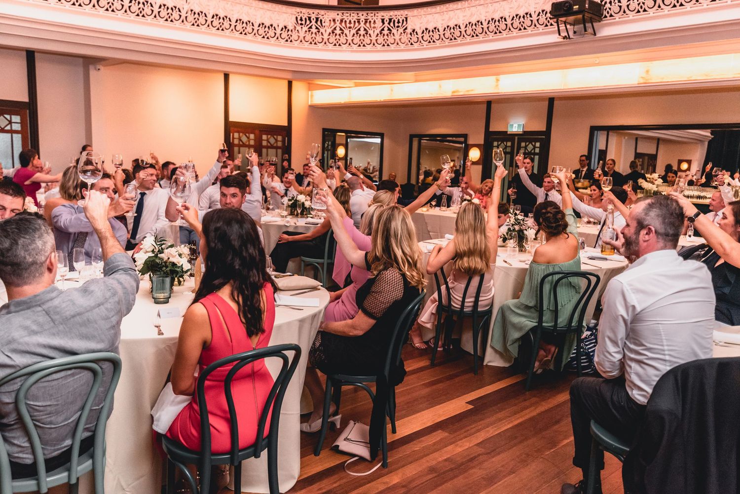 Wedding reception guests seated at round tables in an elegant ballroom with pink lighting and ornate crown molding.
