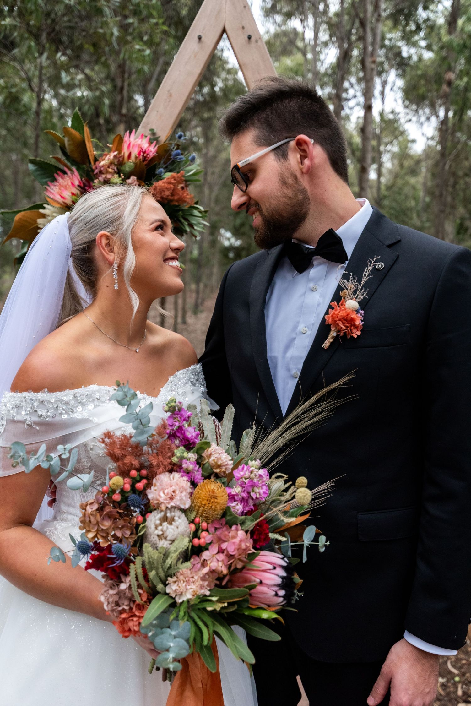 Wedding couple poses with vibrant autumn-colored bouquet against wooden arch backdrop.
